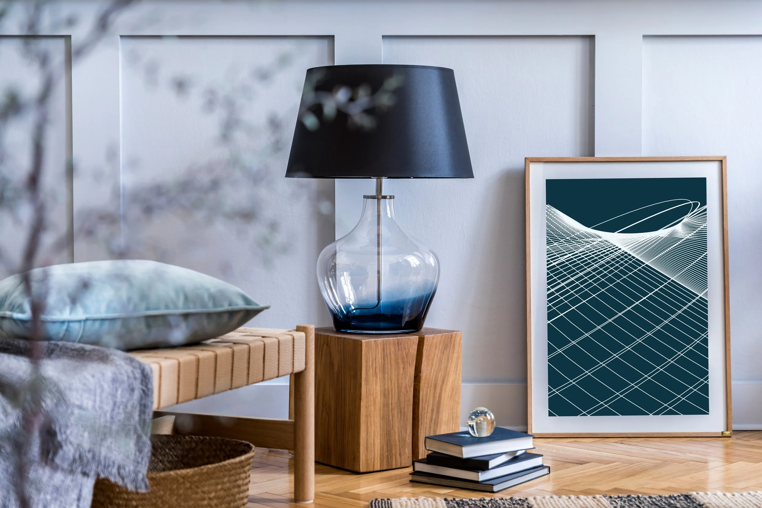 Living room corner with a wooden bench, a blue pillow, a wicker basket, a wooden side table with a glass lamp with a black shade, a stack of books, a glass paperweight, and framed abstract blue and white artwork leaning against the wall.