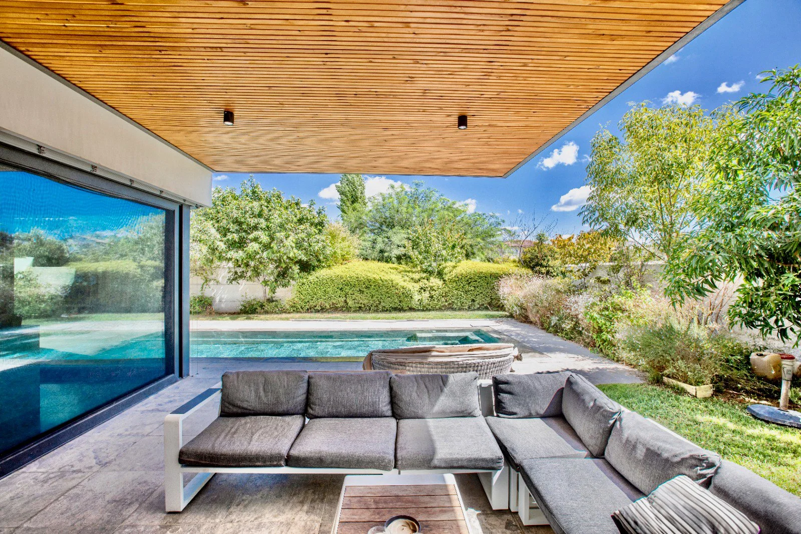 Outdoor patio with gray sectional sofa, view of backyard with pool, landscaped garden, wooden ceiling, and blue sky with clouds.