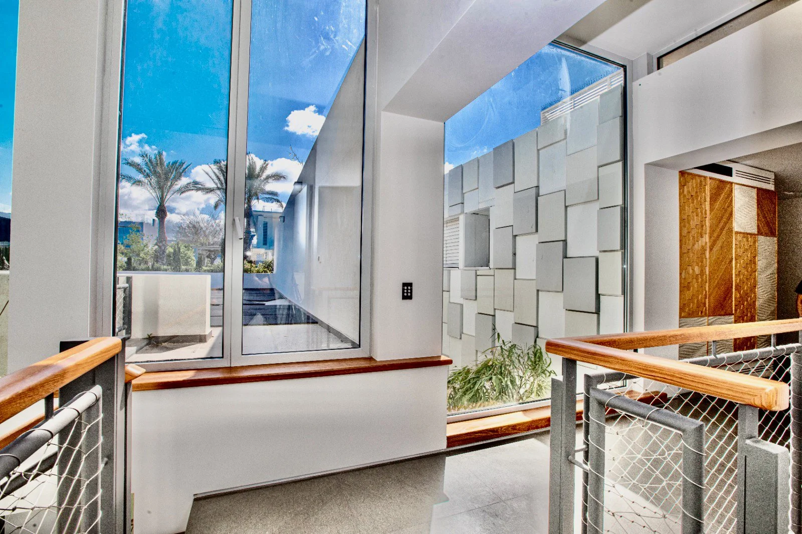 Interior shot of a modern building with large glass windows showing palm trees and blue sky outside. The space features a wooden handrail and geometric wall design with white panels. Sunlight illuminates the area.