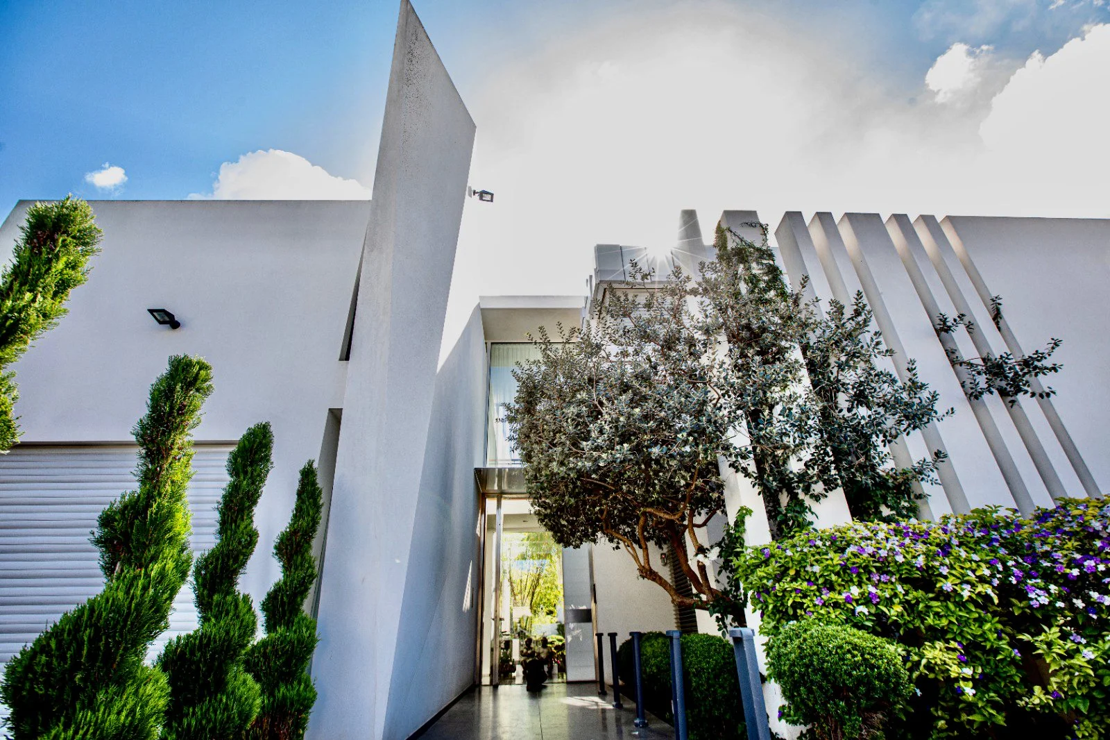 Modern white building with vertical architectural elements and lush green plants, including a tree with purple flowers, under a partly cloudy sky.