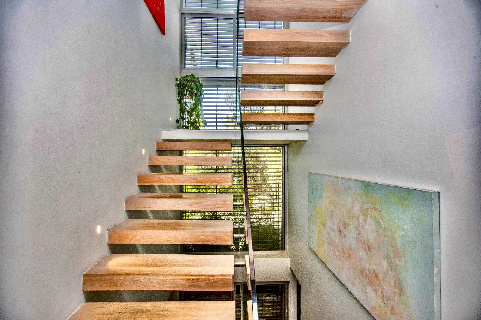 Modern interior space featuring a staircase with wooden steps and a black handrail, large windows with horizontal blinds, a plant, a colorful abstract painting, and a gray wall.