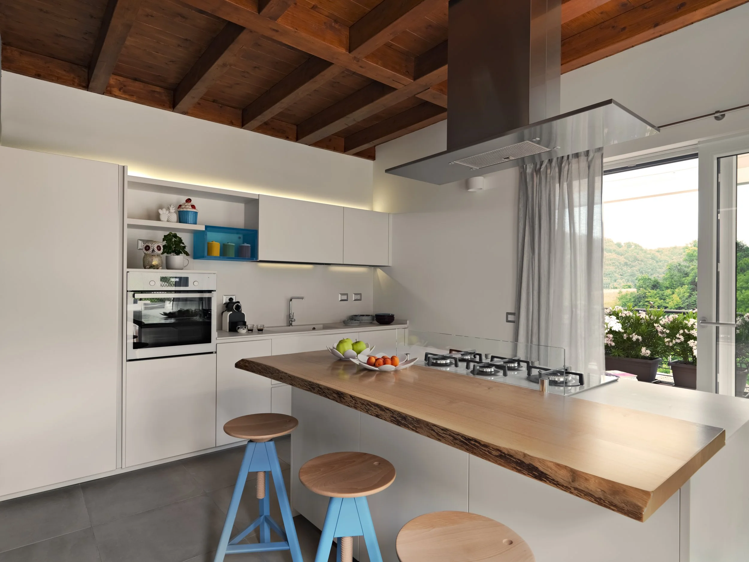 Modern kitchen with white cabinetry, a wooden countertop island, two blue stools, and a view of a balcony with potted flowers and greenery outside.
