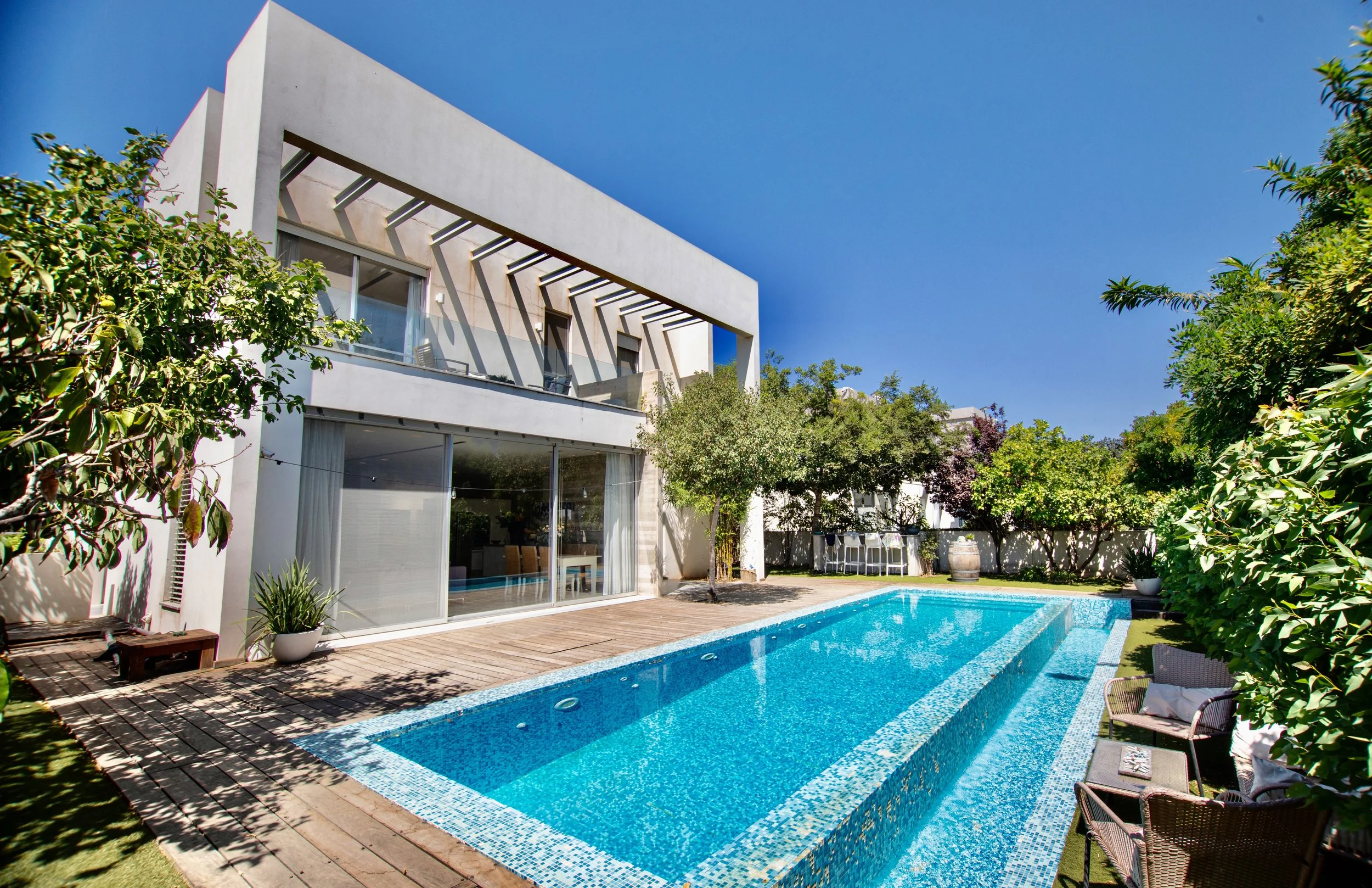Modern white house with large glass sliding doors, outdoor pool, wooden deck, and lush greenery under a clear blue sky.