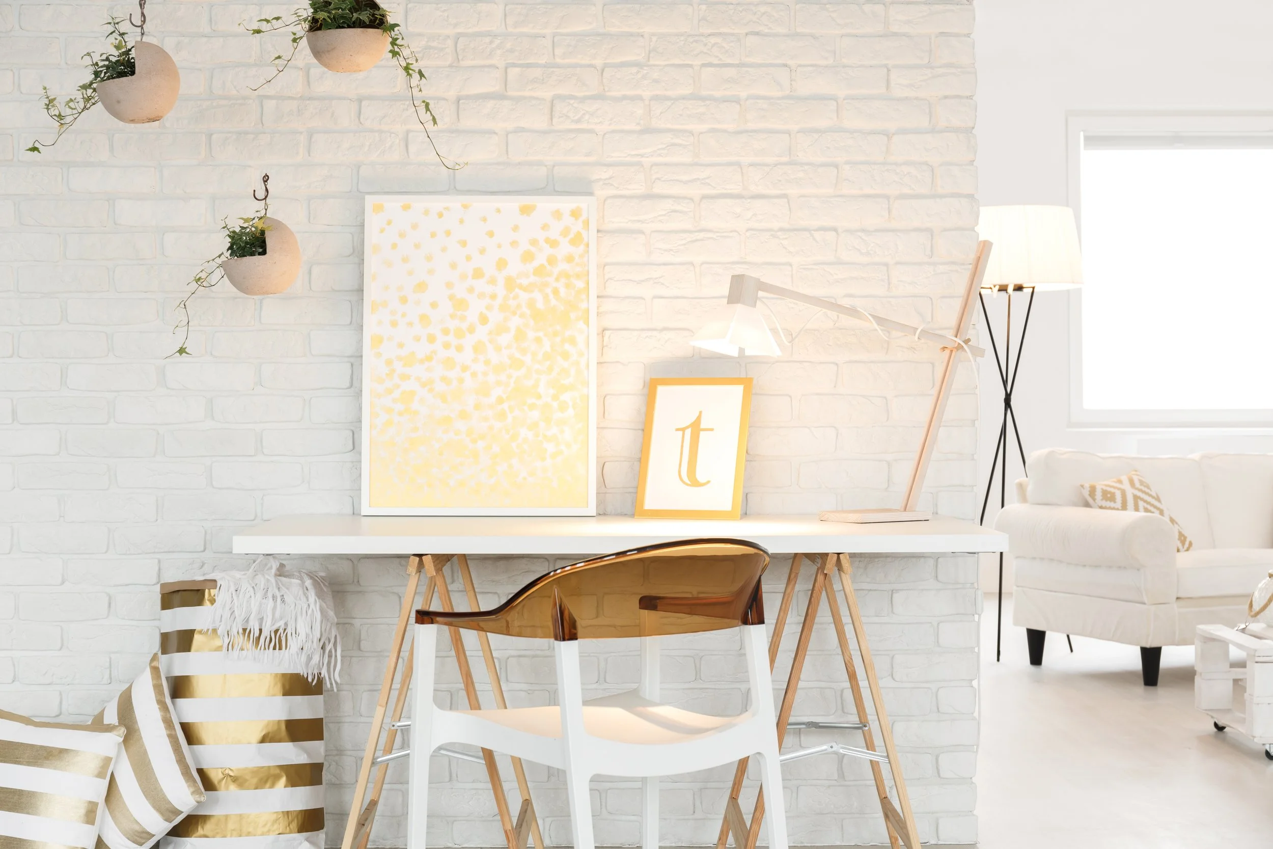 Bright interior with white brick wall, white table, a chair with a wooden back and metal legs, framed artwork with yellow and white pattern, a large framed yellow letter 't', white sofa, lamp, hanging planters, and decorative pillows.