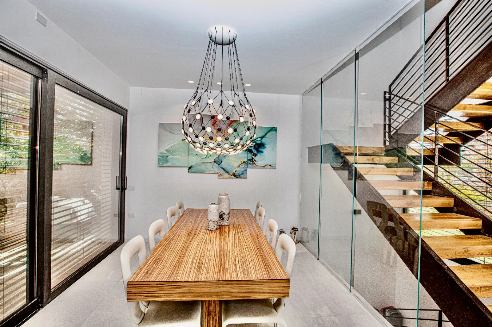 Modern dining room with a wooden table, white chairs, abstract wall art, large chandelier, glass staircase, and sliding glass door.