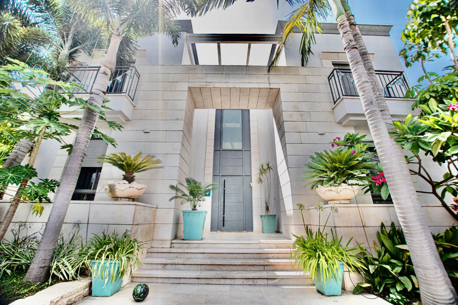 Modern multi-story building with a beige stone facade, large glass door, and a set of stairs leading up to the entrance, surrounded by tropical plants and palm trees.