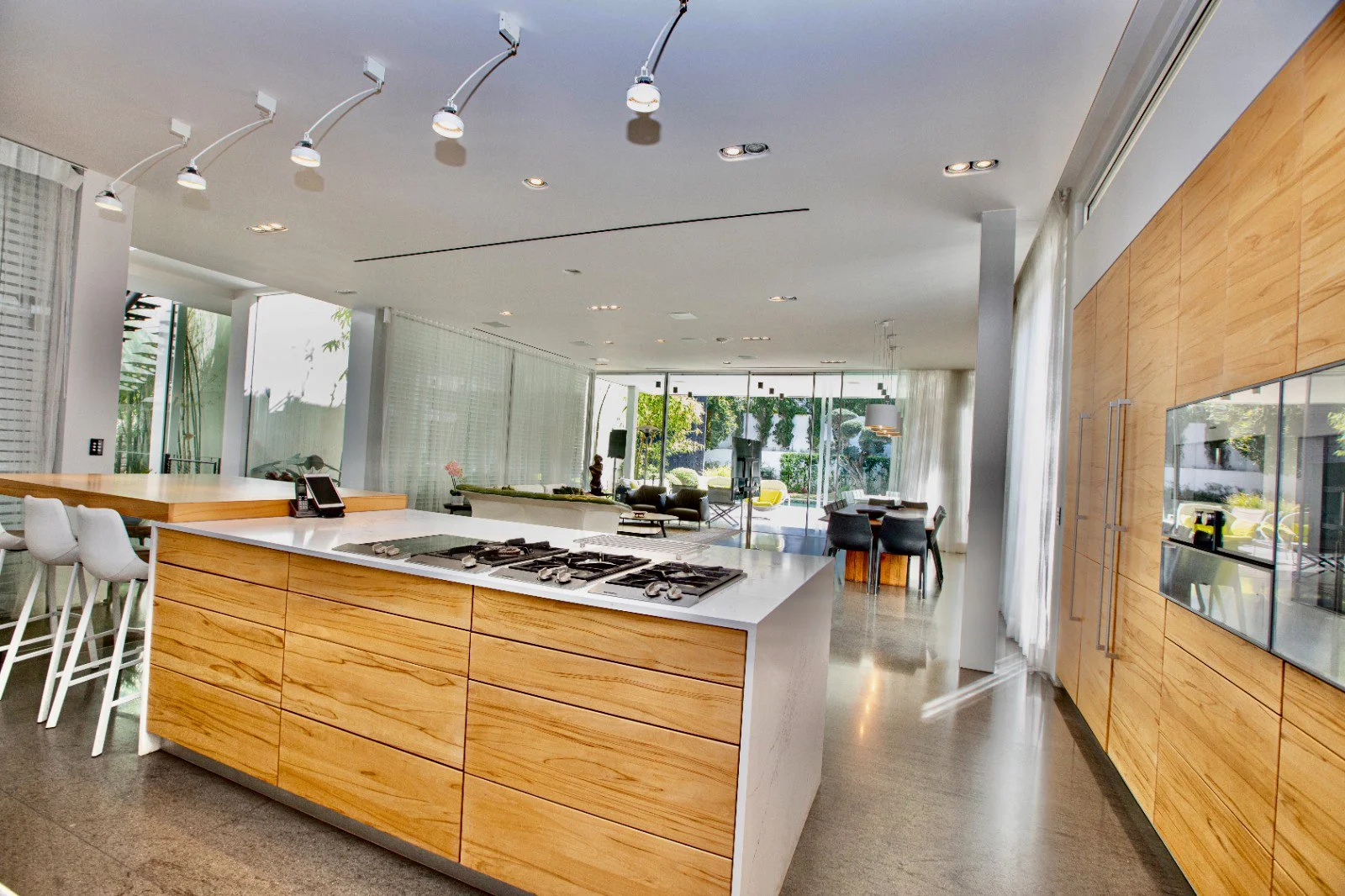 Modern open-concept kitchen with wooden cabinets, white island with built-in gas stove, barstools, and a living area with large windows and outdoor view.
