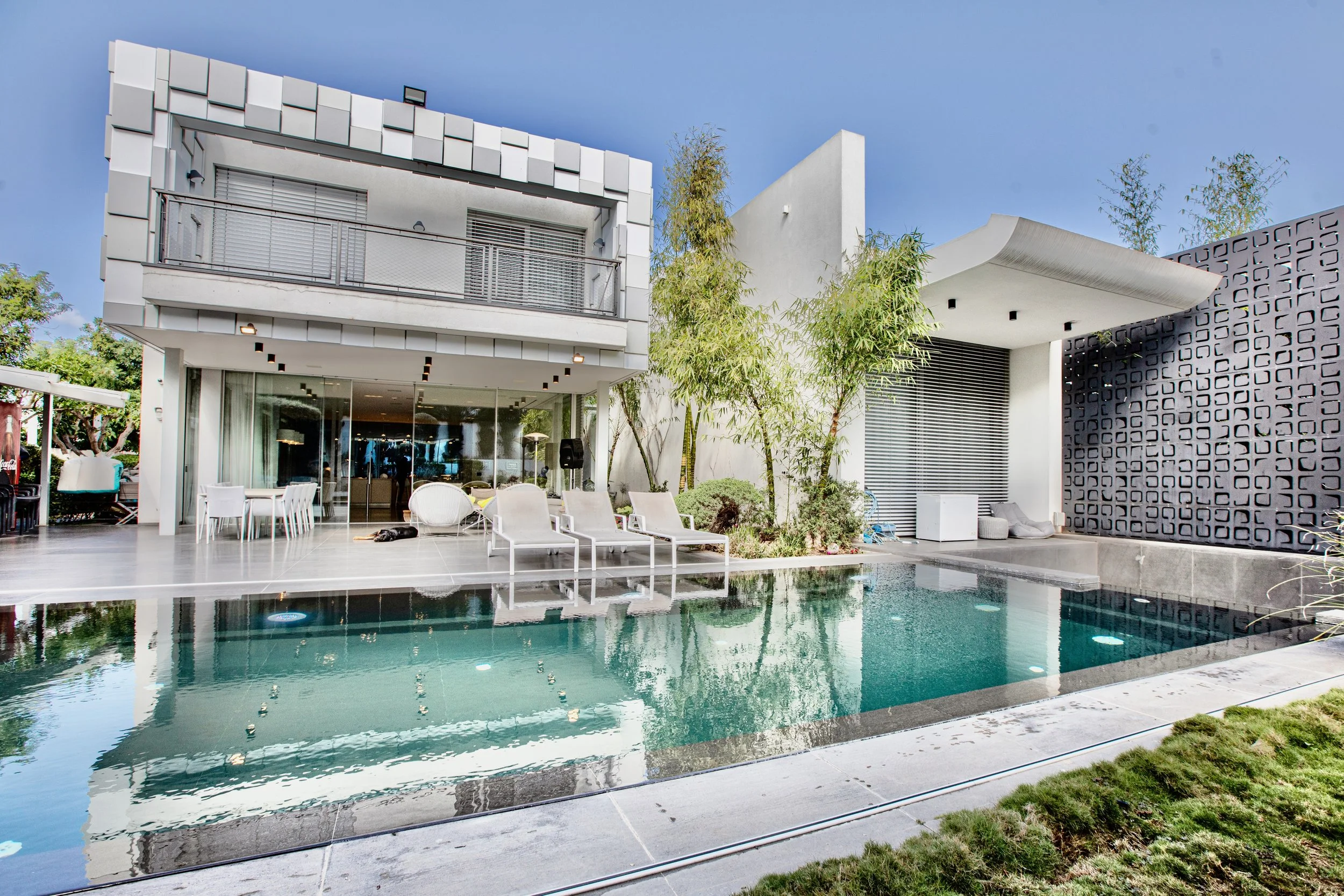 Modern house with swimming pool, outdoor lounge chairs, and patio furniture in backyard, with trees and a clear blue sky.