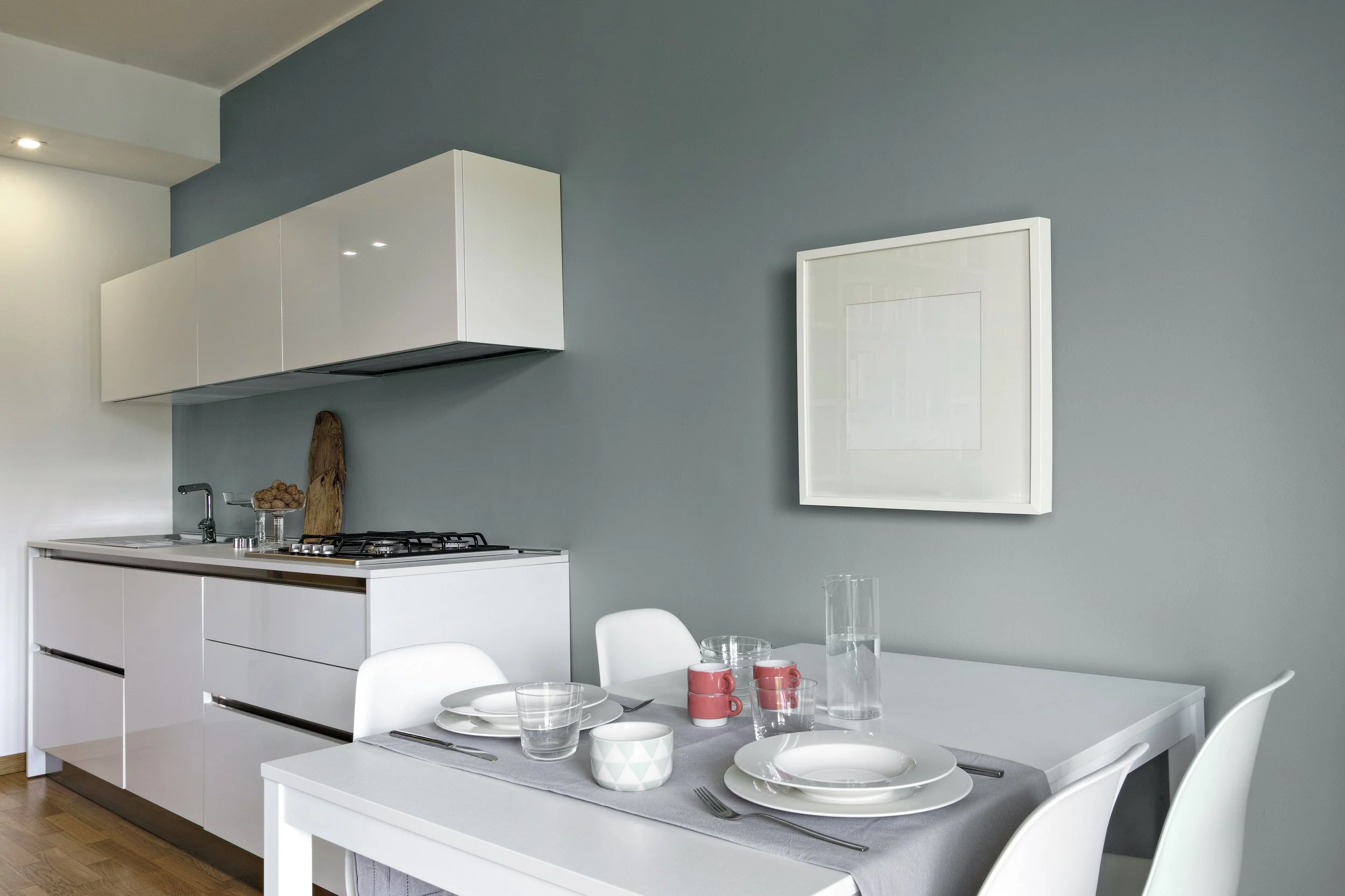Modern kitchen with white cabinets, a gray wall, and a dining table set with white dishes, glasses, and cups.