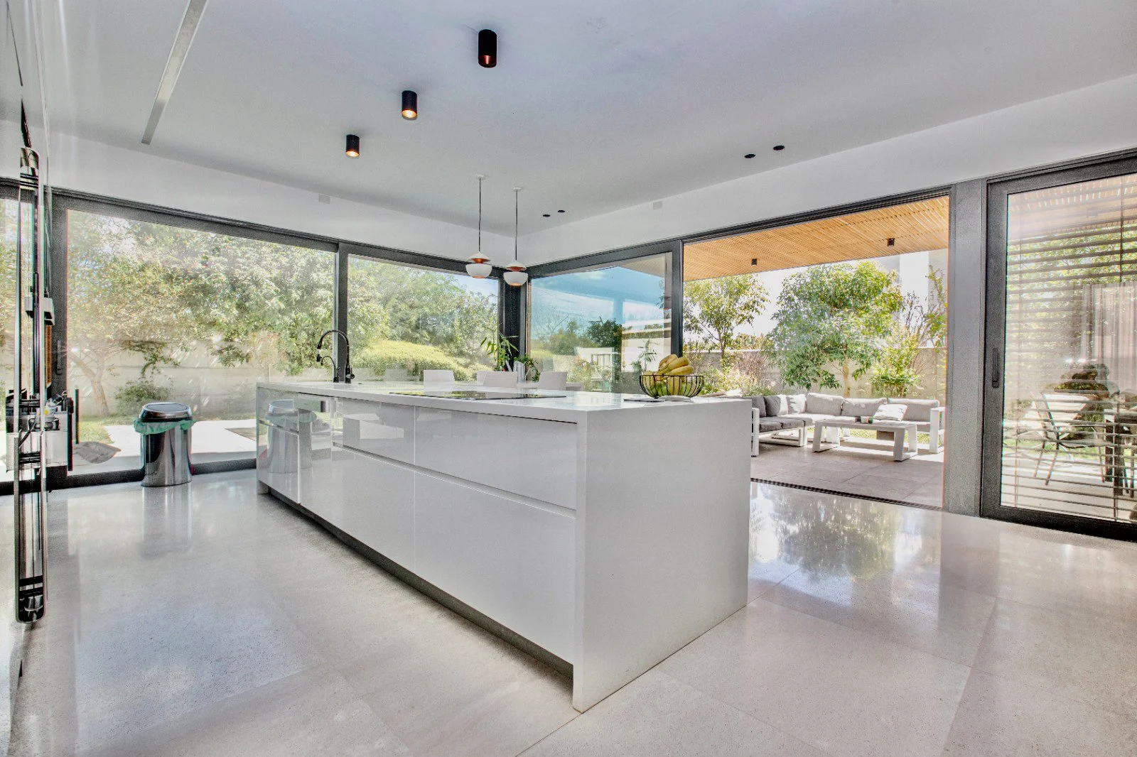 Modern kitchen with white island, large glass sliding doors leading to outdoor patio with seating area, green trees, and a clear sky.