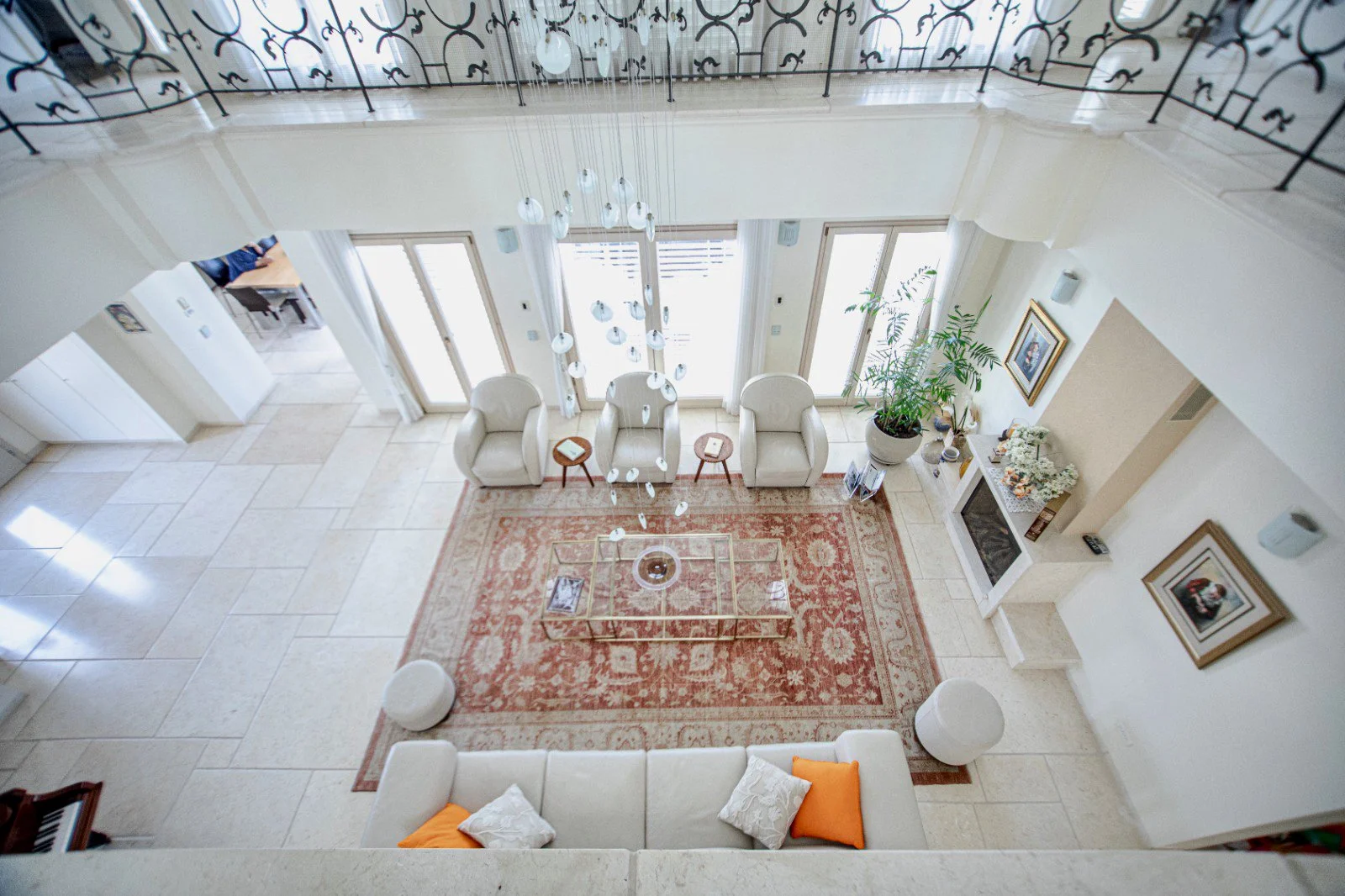 View from above of a bright living room with white chairs, a patterned rug, a glass coffee table, a fireplace, framed photos, potted plants, and glass doors leading to an outdoor balcony.