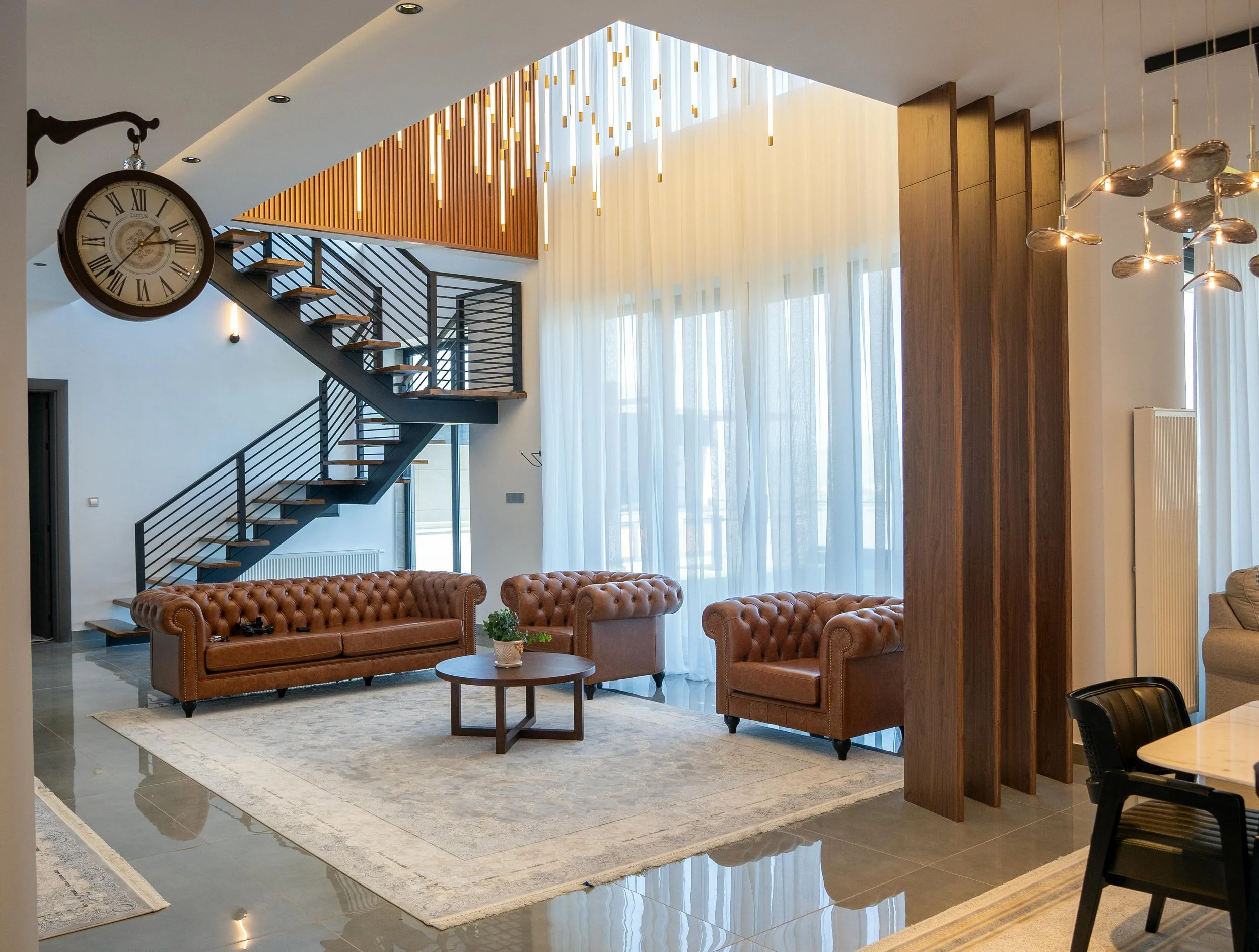 Modern living room with brown leather sofas, a round coffee table with a plant, large windows with sheer curtains, a staircase with black railings, a clock on the wall, and wooden decorative panels.