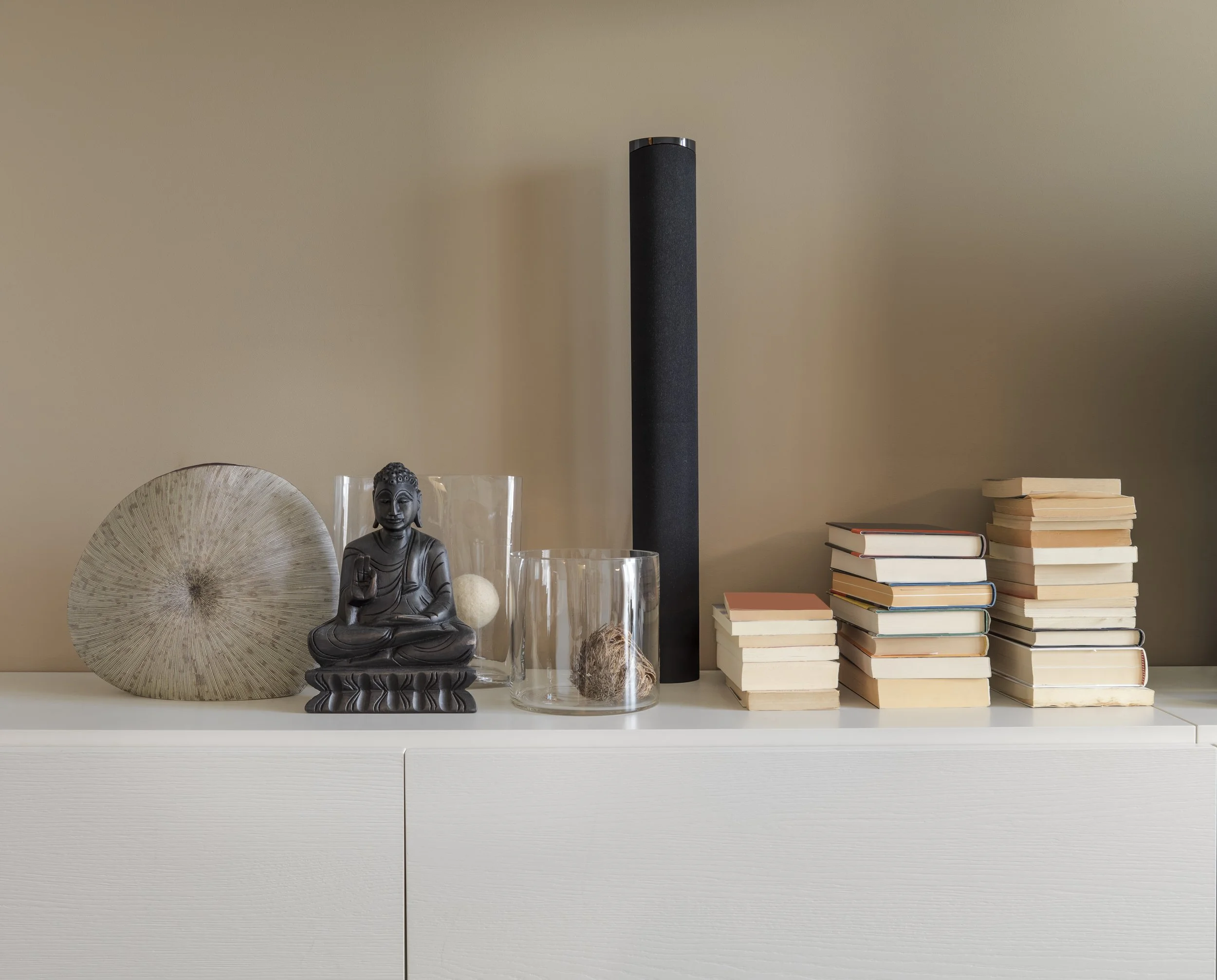 Decorative objects including a large circular textured piece, a black Buddha figurine, glass vases with fibrous objects inside, and stacks of books on a white surface against a beige wall.