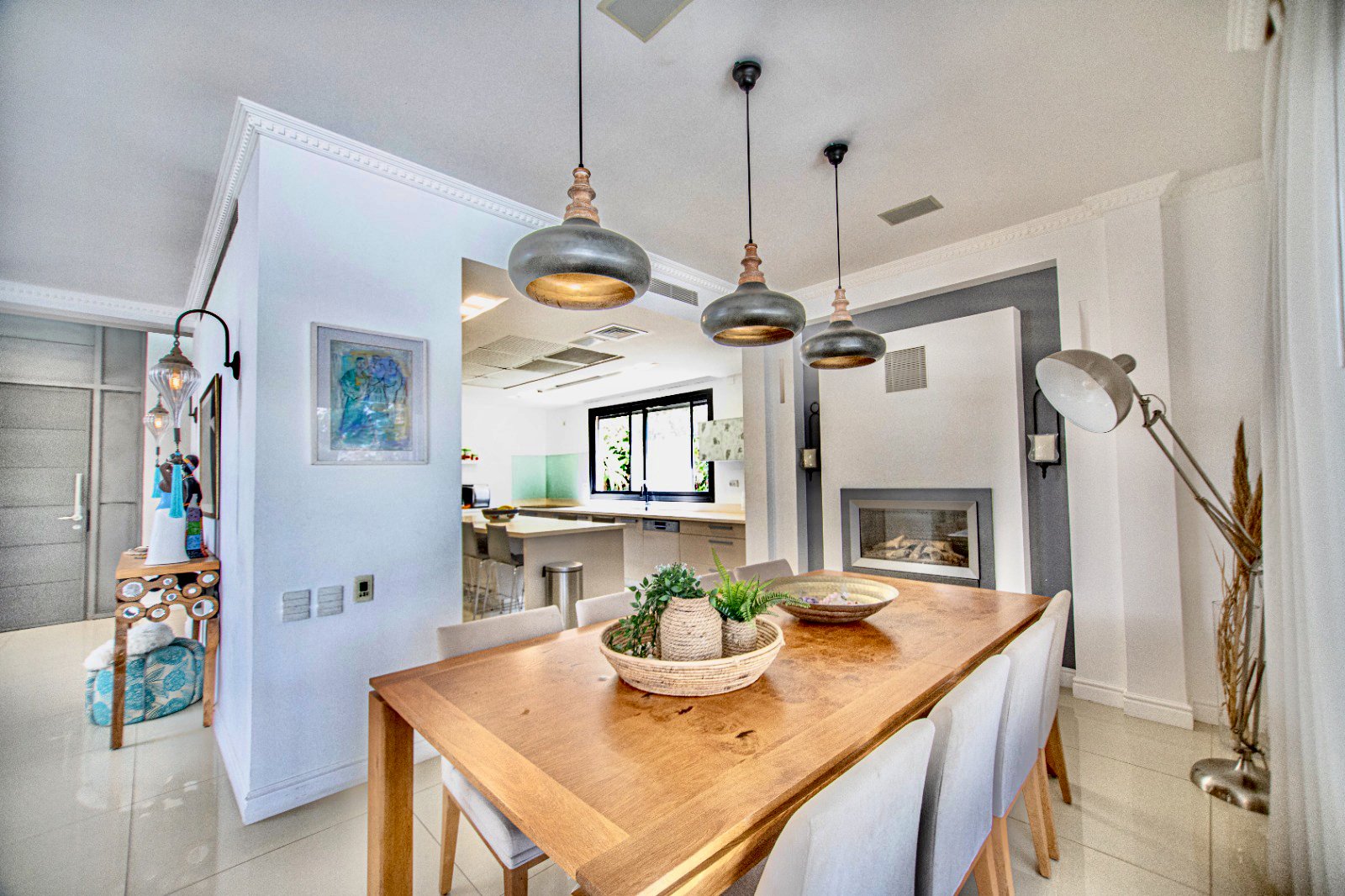 Modern dining room with a wooden table, white chairs, hanging pendant lights, a fireplace, and a large window in background.