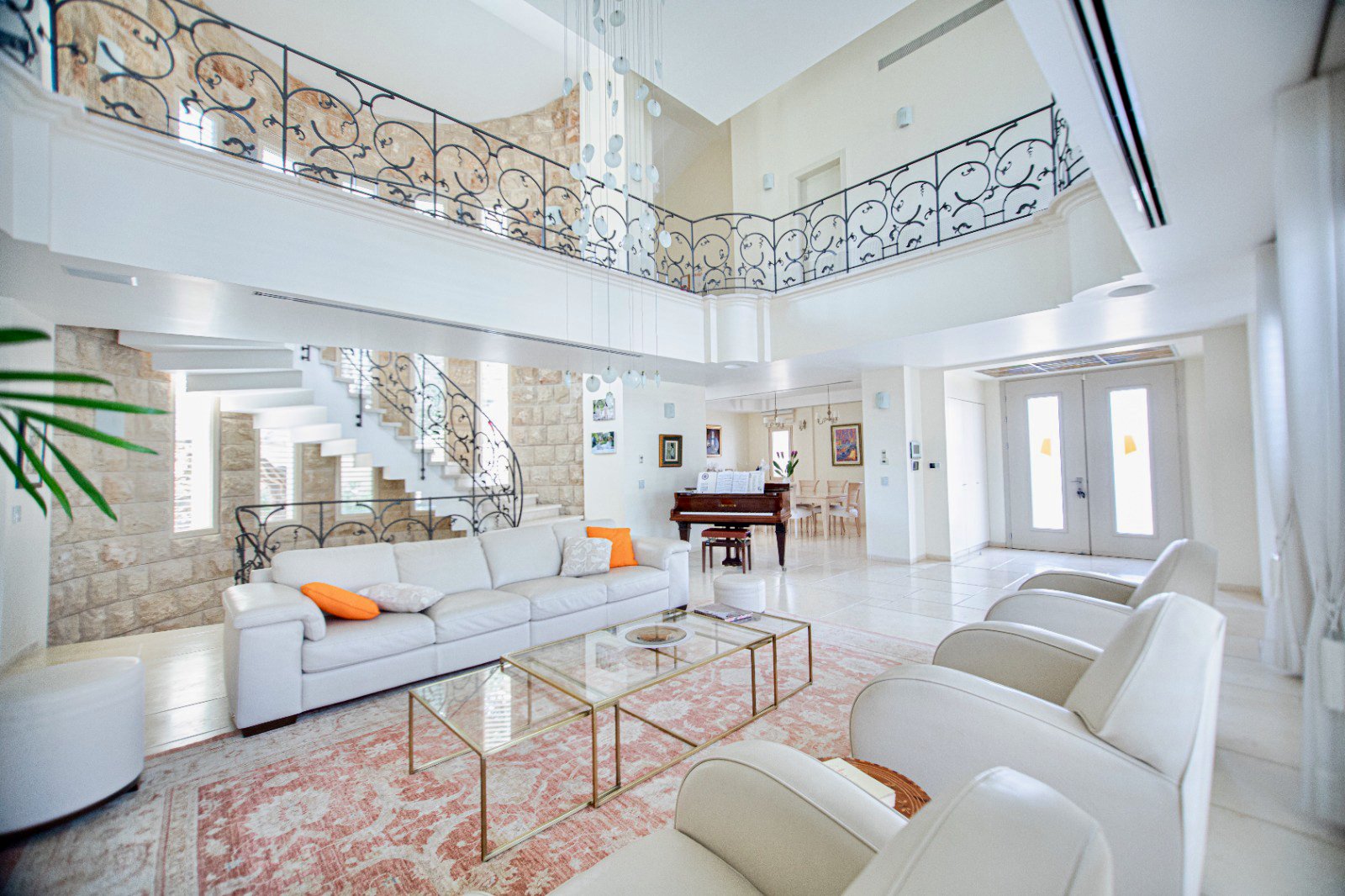 Bright, spacious living room with white sofas, beige armchairs, and a glass coffee table on a pink patterned rug. A grand staircase with ornate black railing leads to the upper level, and there is a piano in the background near a small dining area. D