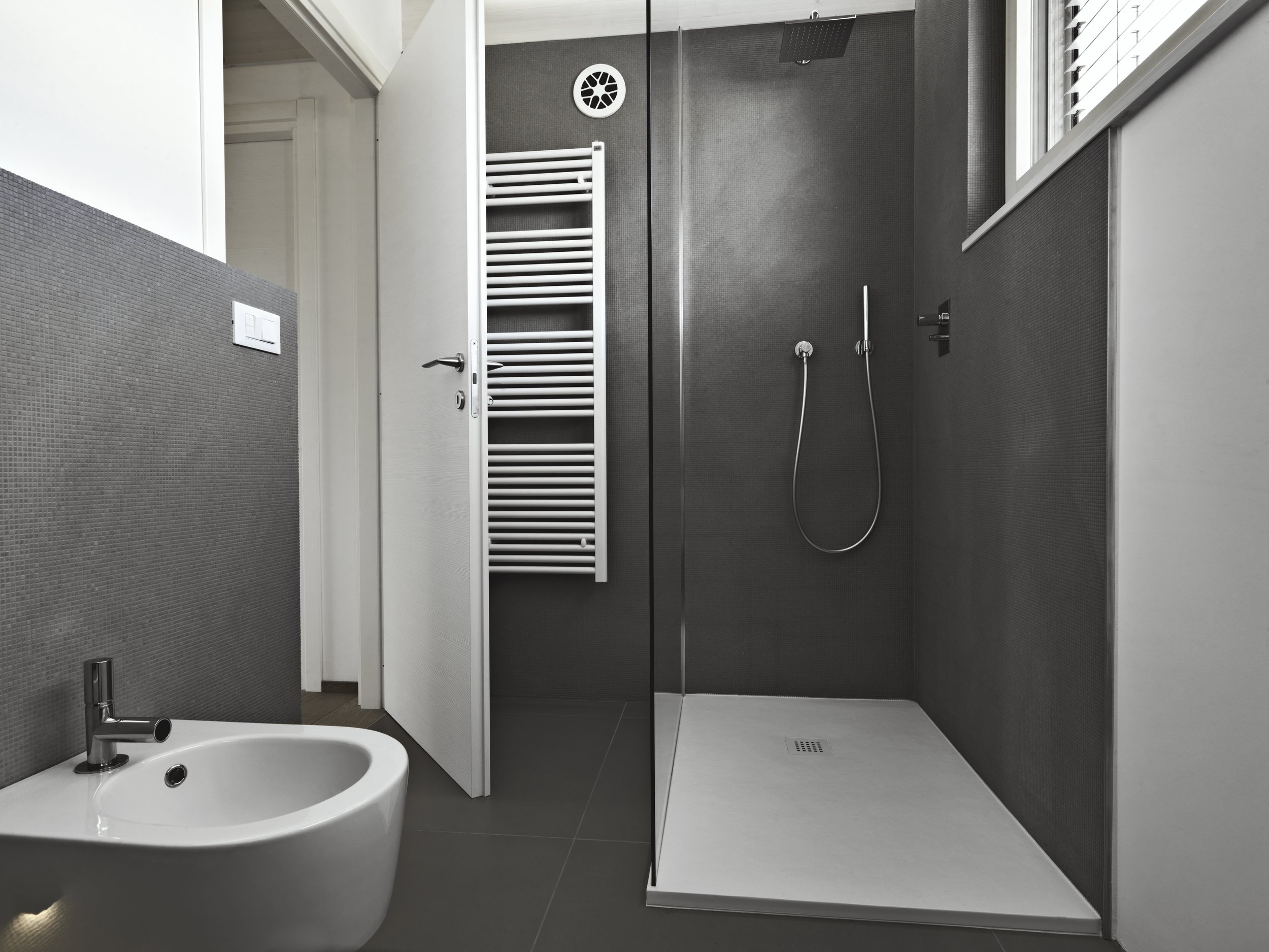 Modern bathroom with a bidet, gray textured wall, white door, towel radiator, shower area with handheld showerhead, large window with blinds.