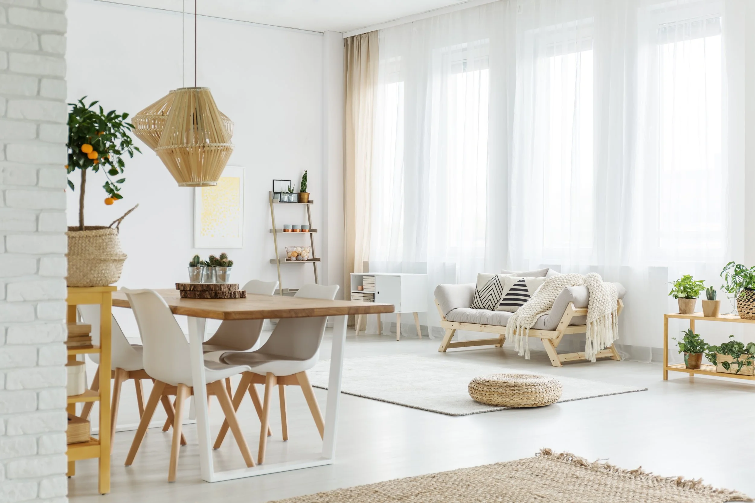 Bright living room with white walls, large windows with sheer curtains, a white sofa with patterned pillows, a wooden coffee table, a woven pouf, green potted plants, a white bookshelf, and a wooden ladder shelf with decorative items.