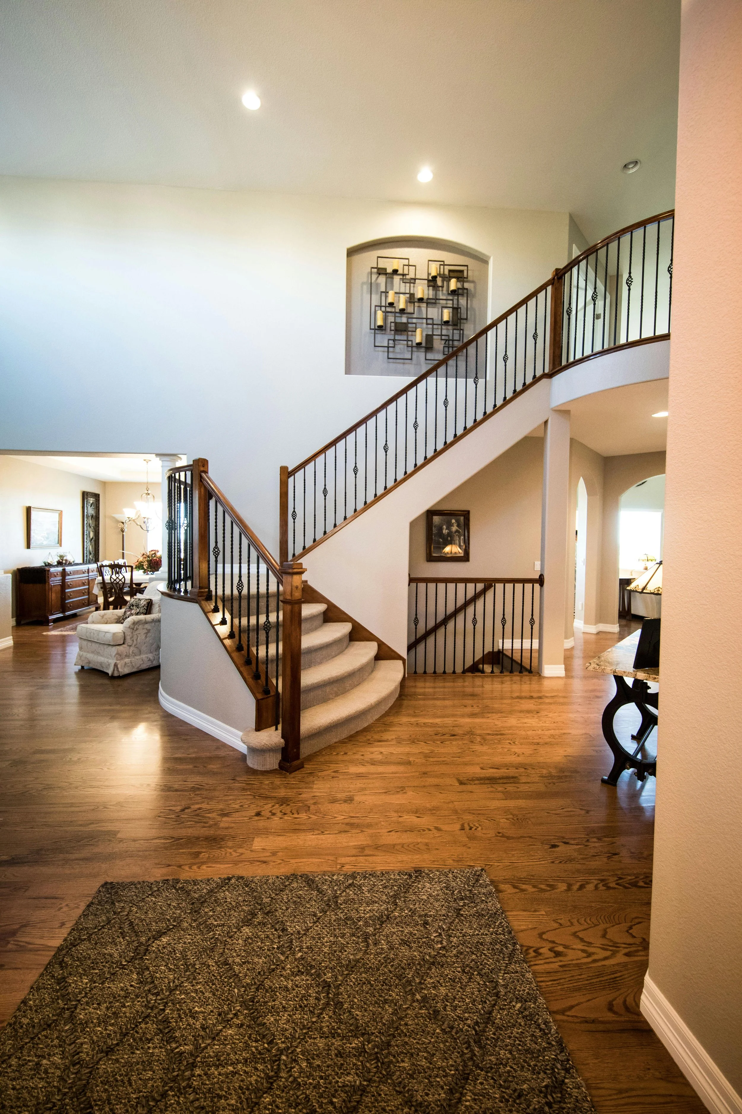 Interior of a spacious home with a curved staircase, hardwood floors, and a decorative wall piece with candles. The entrance leads to a living room with furniture and a dining area in the background.