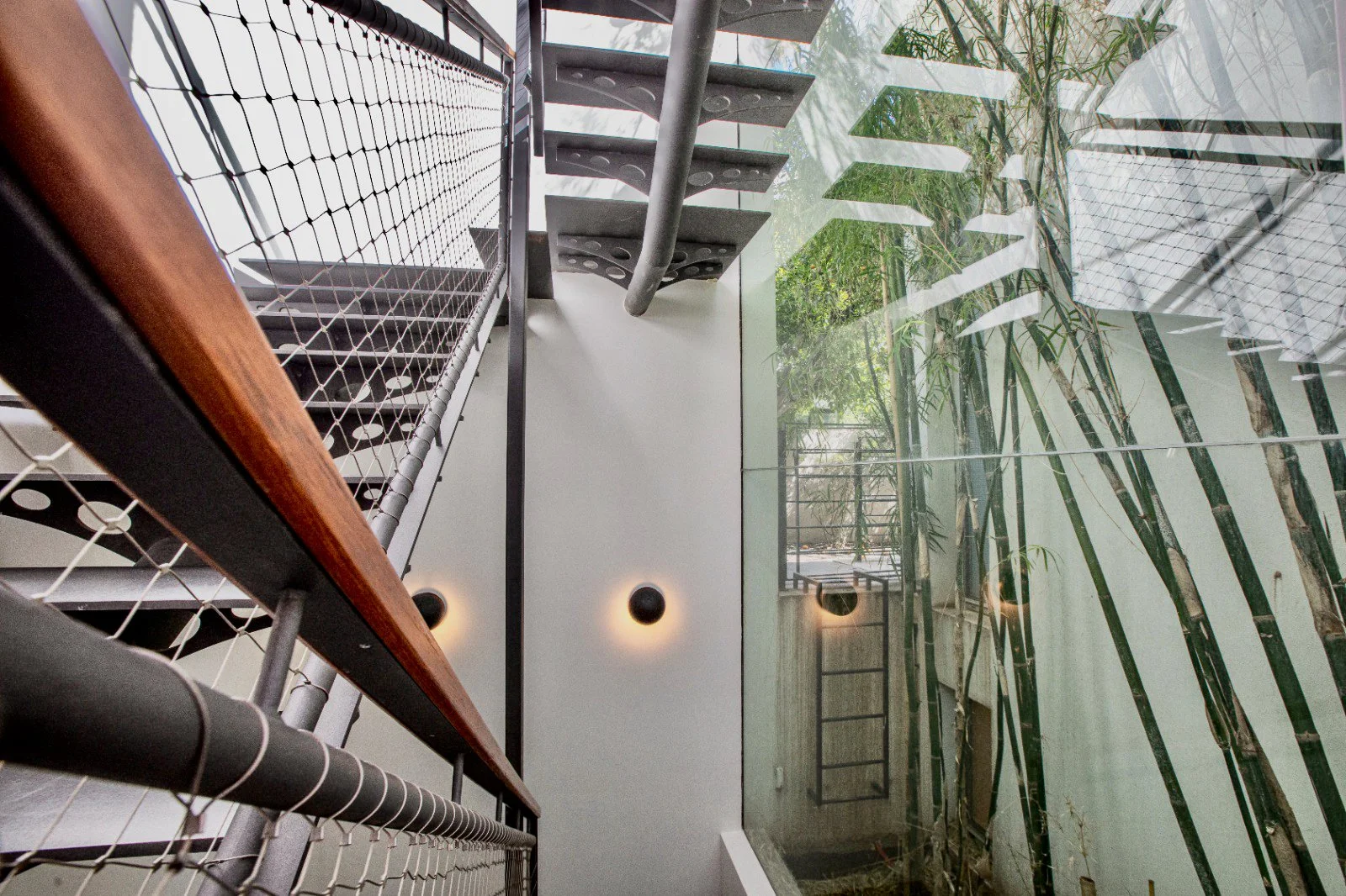 Interior view looking up at stairwell with metal stairs, wooden railings, glass wall with bamboo plants outside, and modern wall lights.