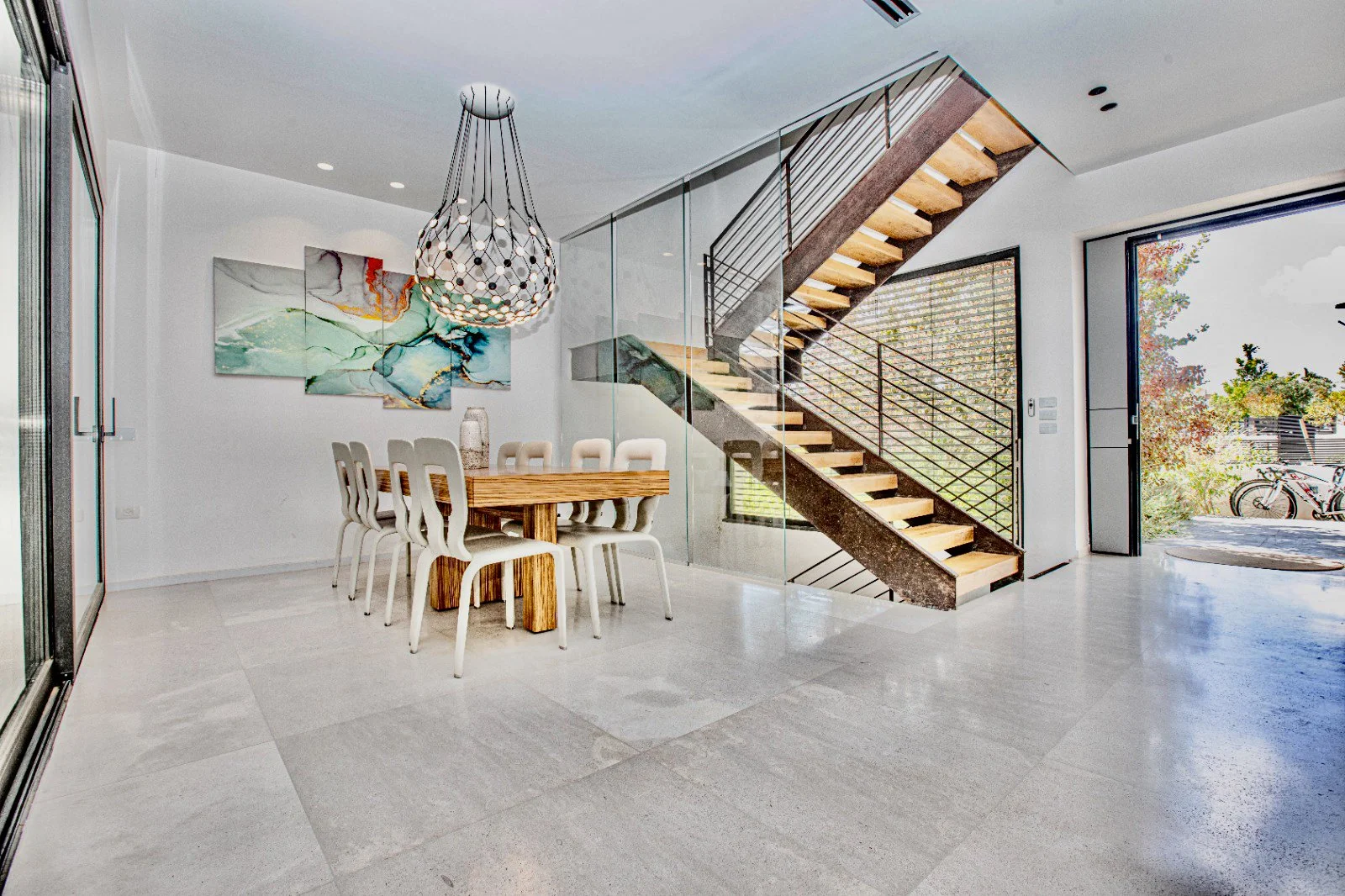 Modern dining area with a wooden table and white chairs, a large multi-colored abstract painting on the white wall, a glass patio door, a staircase with metal railing, and a view of outdoor greenery and bicycles.