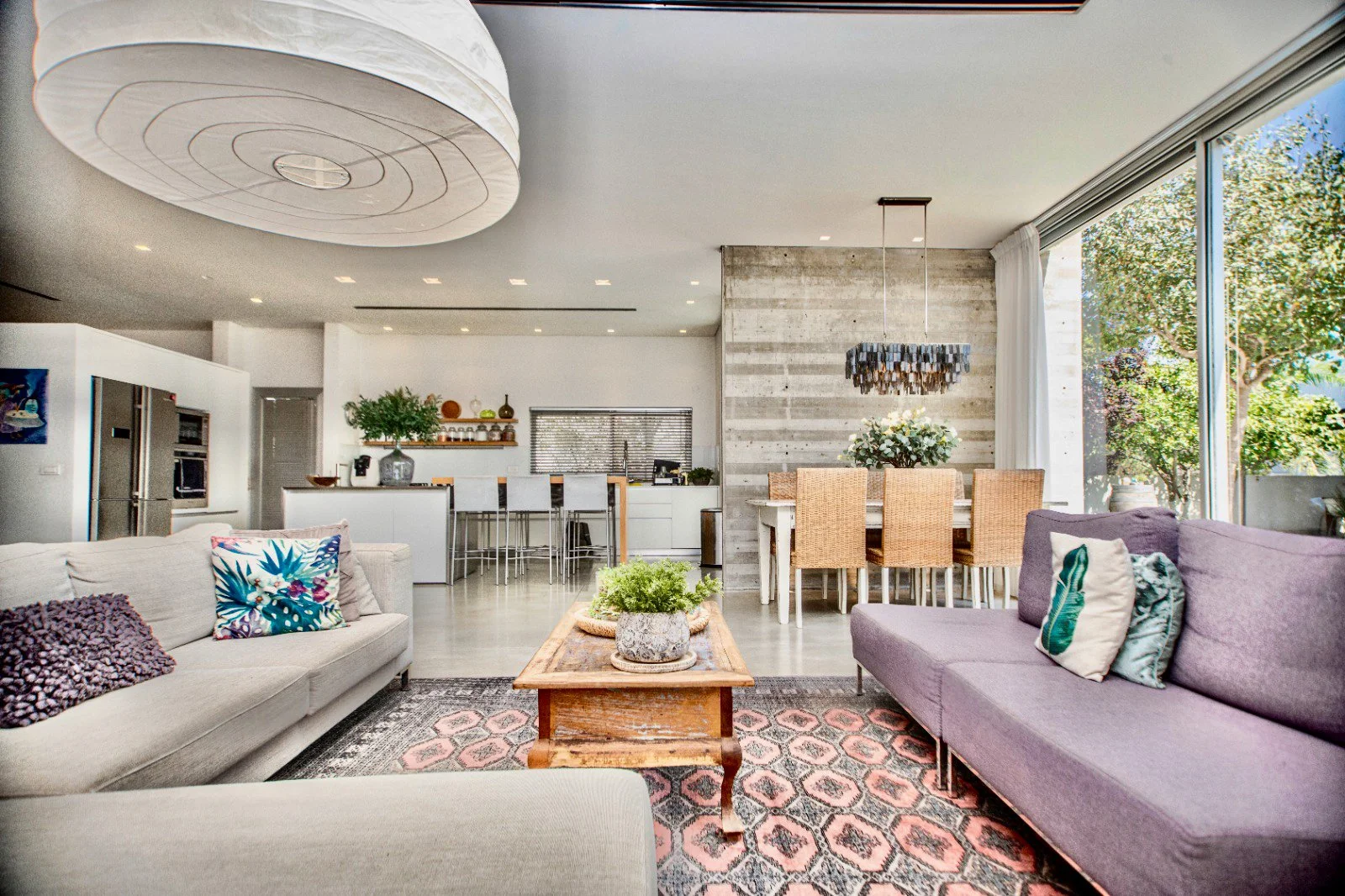 Bright open-concept living room with purple and beige sofas, wooden coffee table, patterned area rug, large window with curtains, and dining area with six woven chairs and a chandelier.
