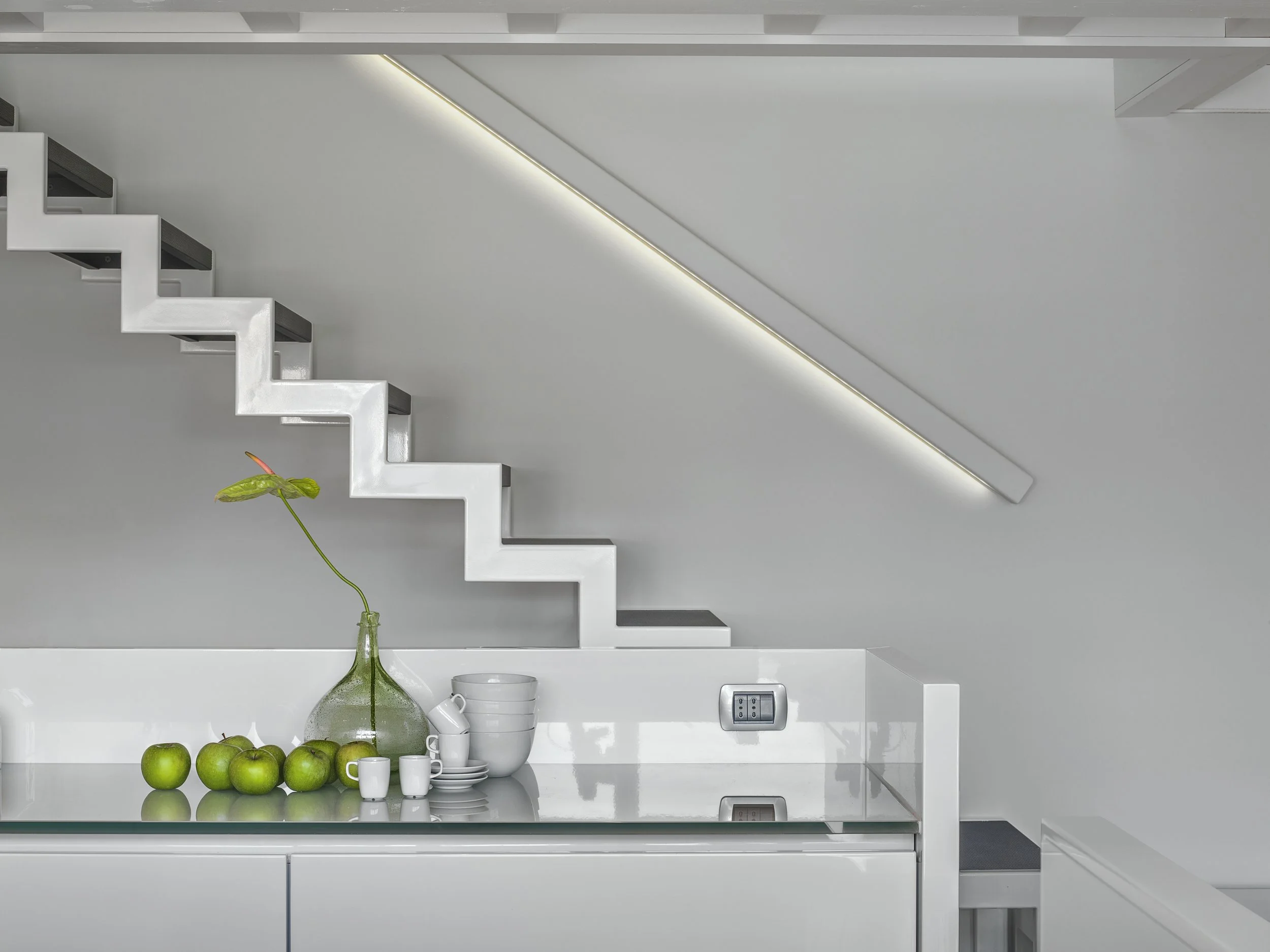 Modern minimalist kitchen with white surfaces, a glass countertop, a green vase with a single leaf, green apples, white cups, and a staircase with zigzag design and integrated lighting.