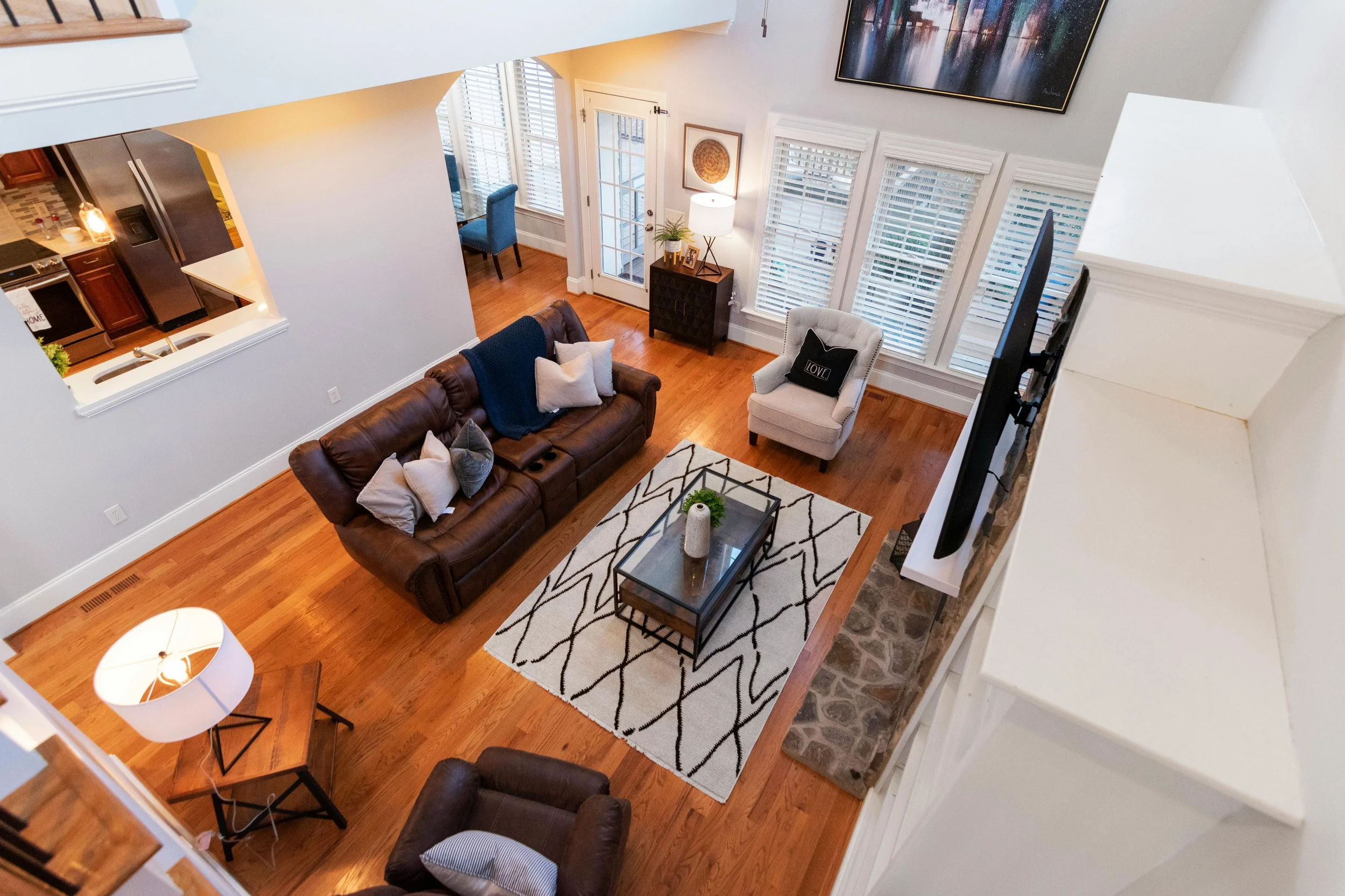 Living room with brown leather sofa, beige armchair, patterned area rug, wooden flooring, and large windows with blinds.