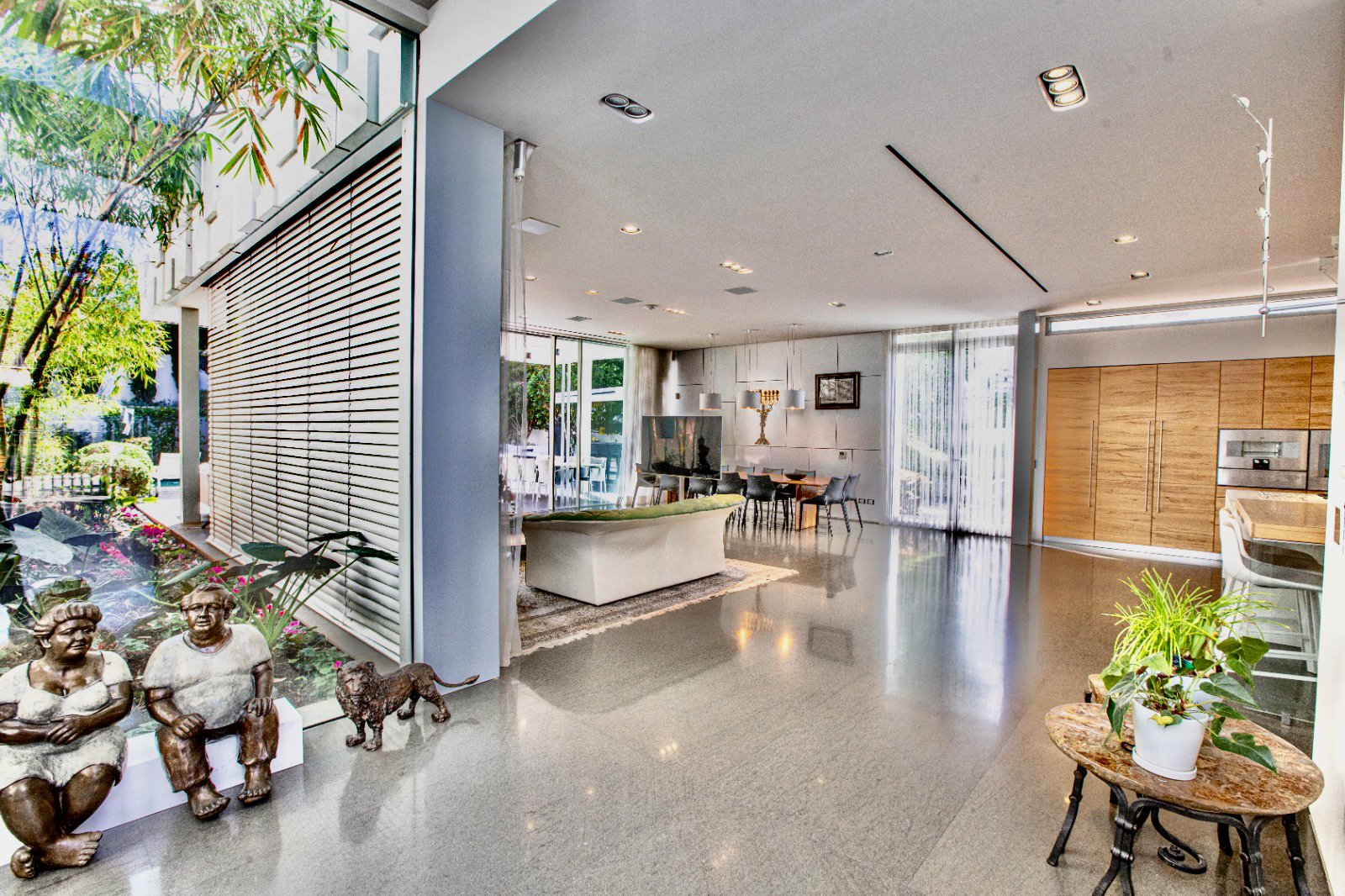 Modern open-concept living room with large glass windows, a seating area with a white sofa, a dining table with chairs, potted plants, and decorative statues near the window