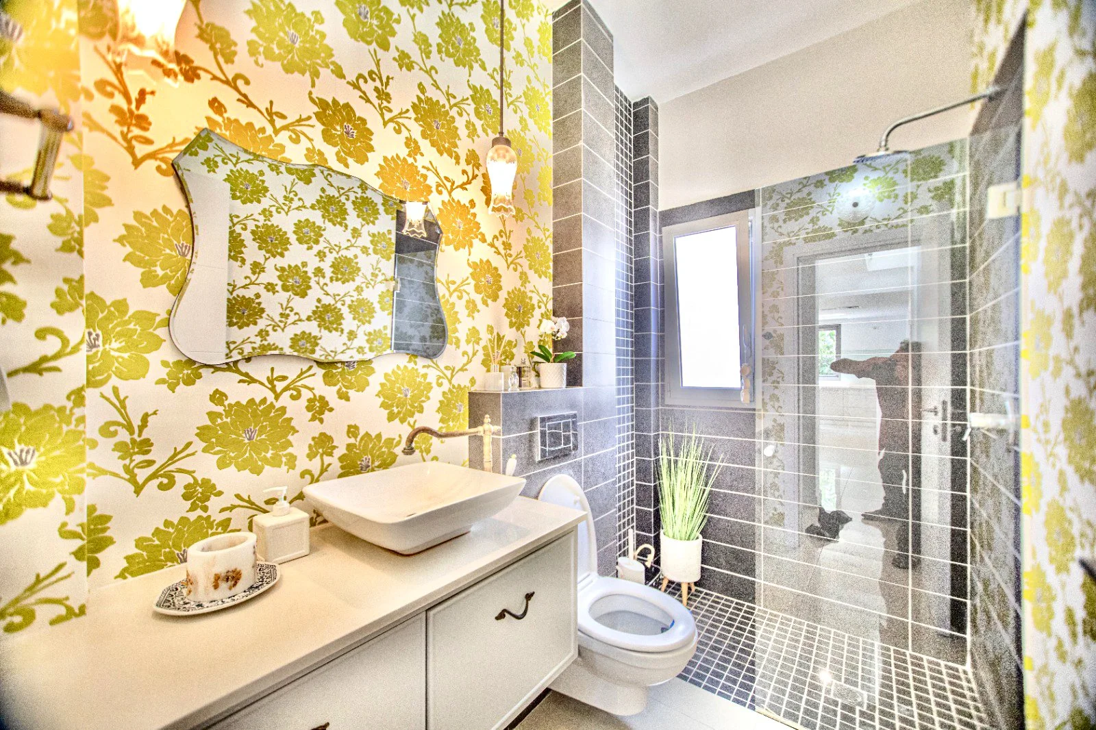 A modern bathroom with yellow floral wallpaper, a white vanity with a rectangular vessel sink, and a purse-shaped mirror. The bathroom features a separate shower area with black and gray tiles, a glass shower door, and a window. There are plants and 