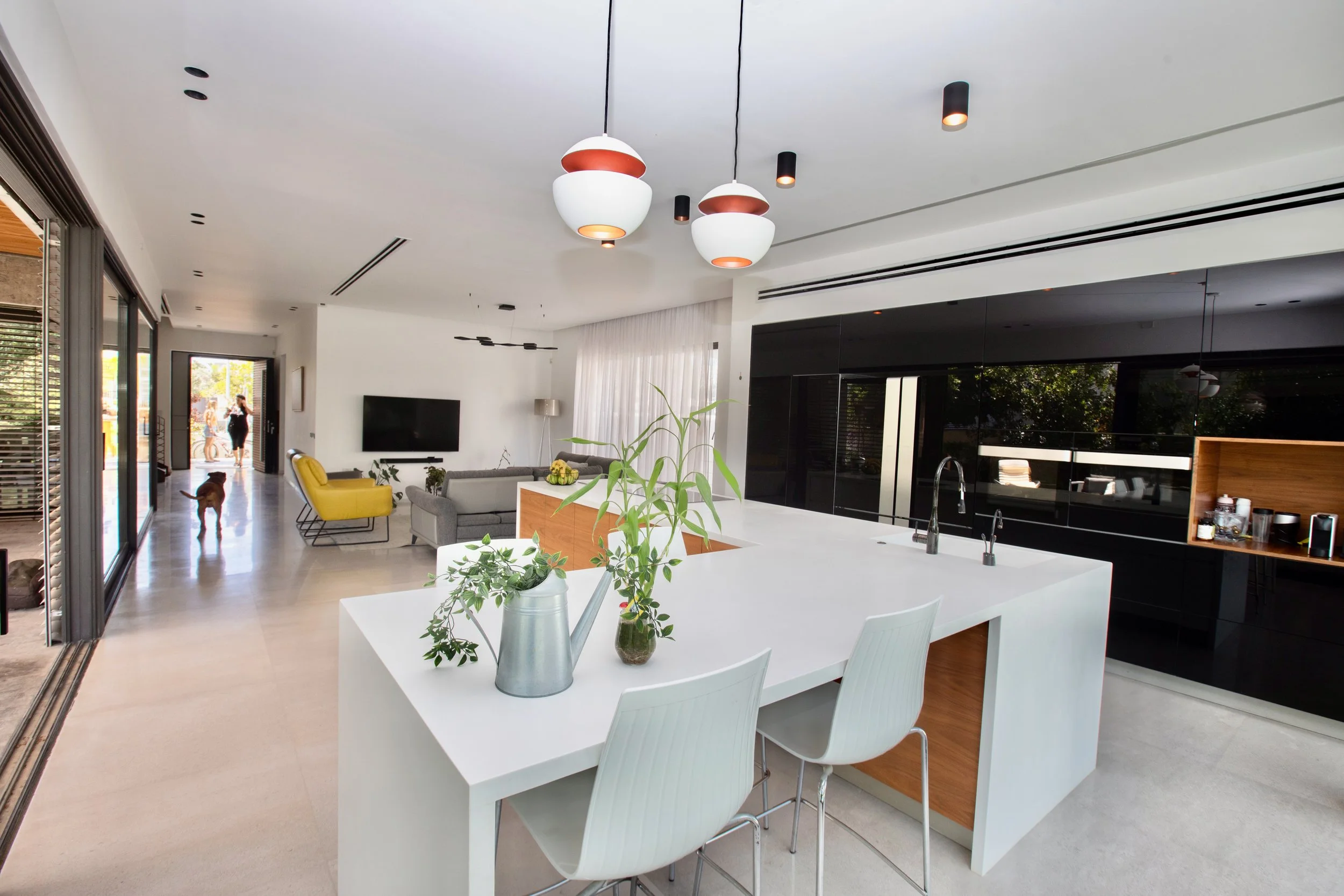 Open-concept modern kitchen with white island, green plants, and black cabinetry, connected to living area with yellow armchair, gray sofa, and wall-mounted TV