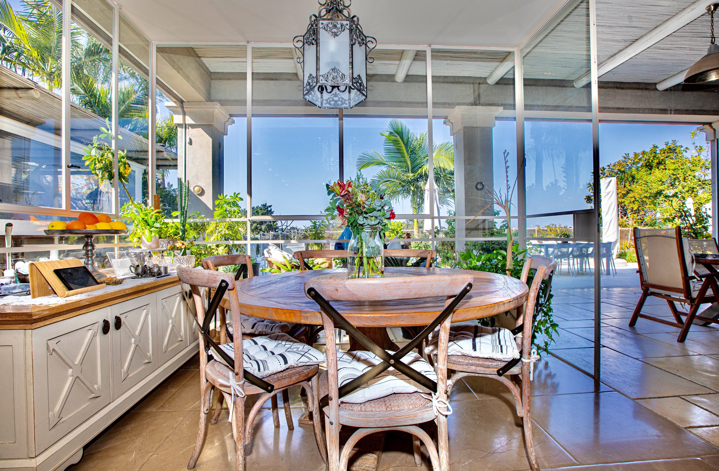 Bright indoor dining area with a round wooden table, surrounded by chairs with cushions, set near large glass windows showing a patio with outdoor furniture and lush green plants including palm trees.