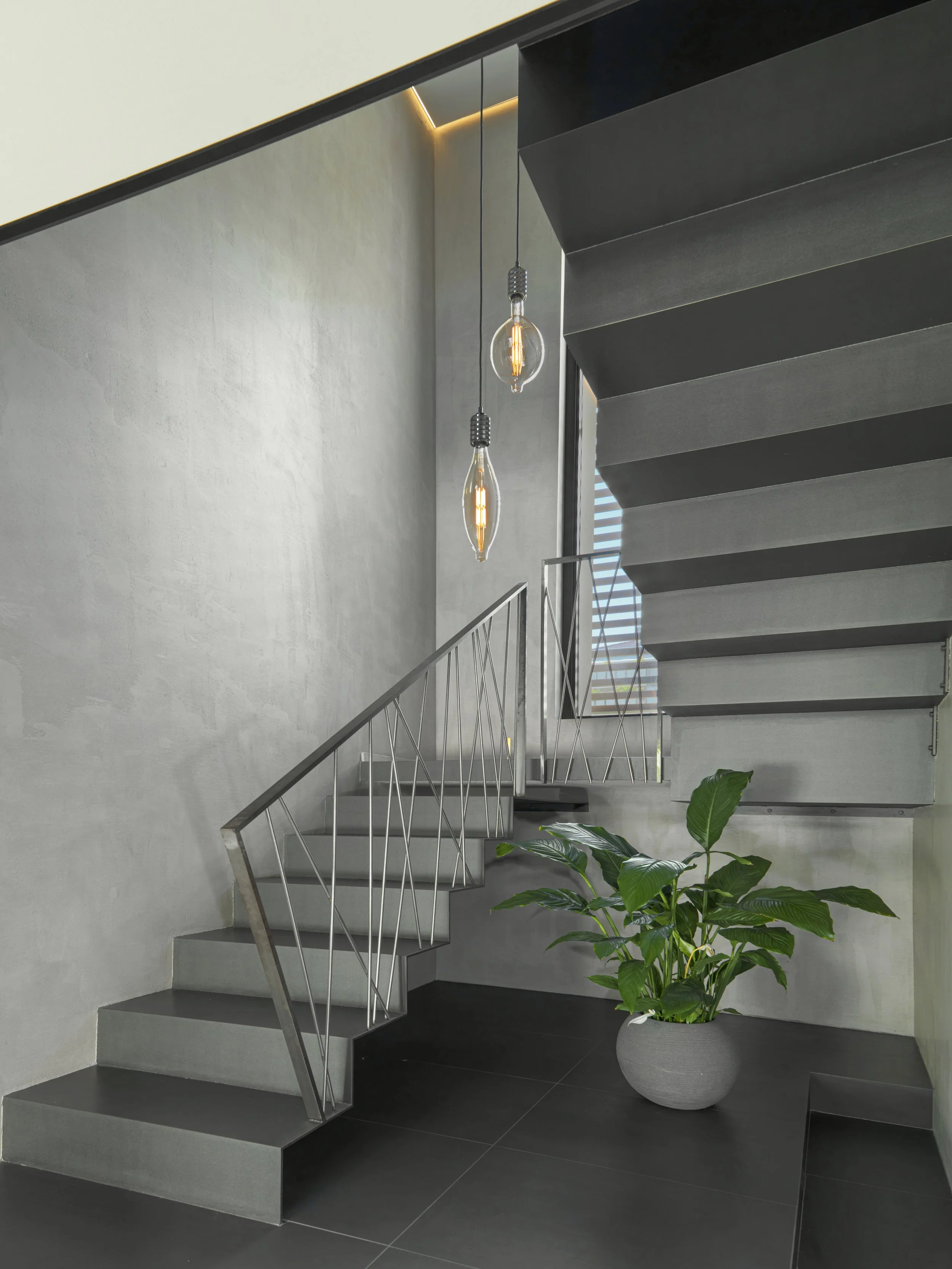 Modern interior staircase with concrete steps, metal railing, hanging Edison-style light bulbs, potted green plant, gray textured walls, and window with horizontal blinds.
