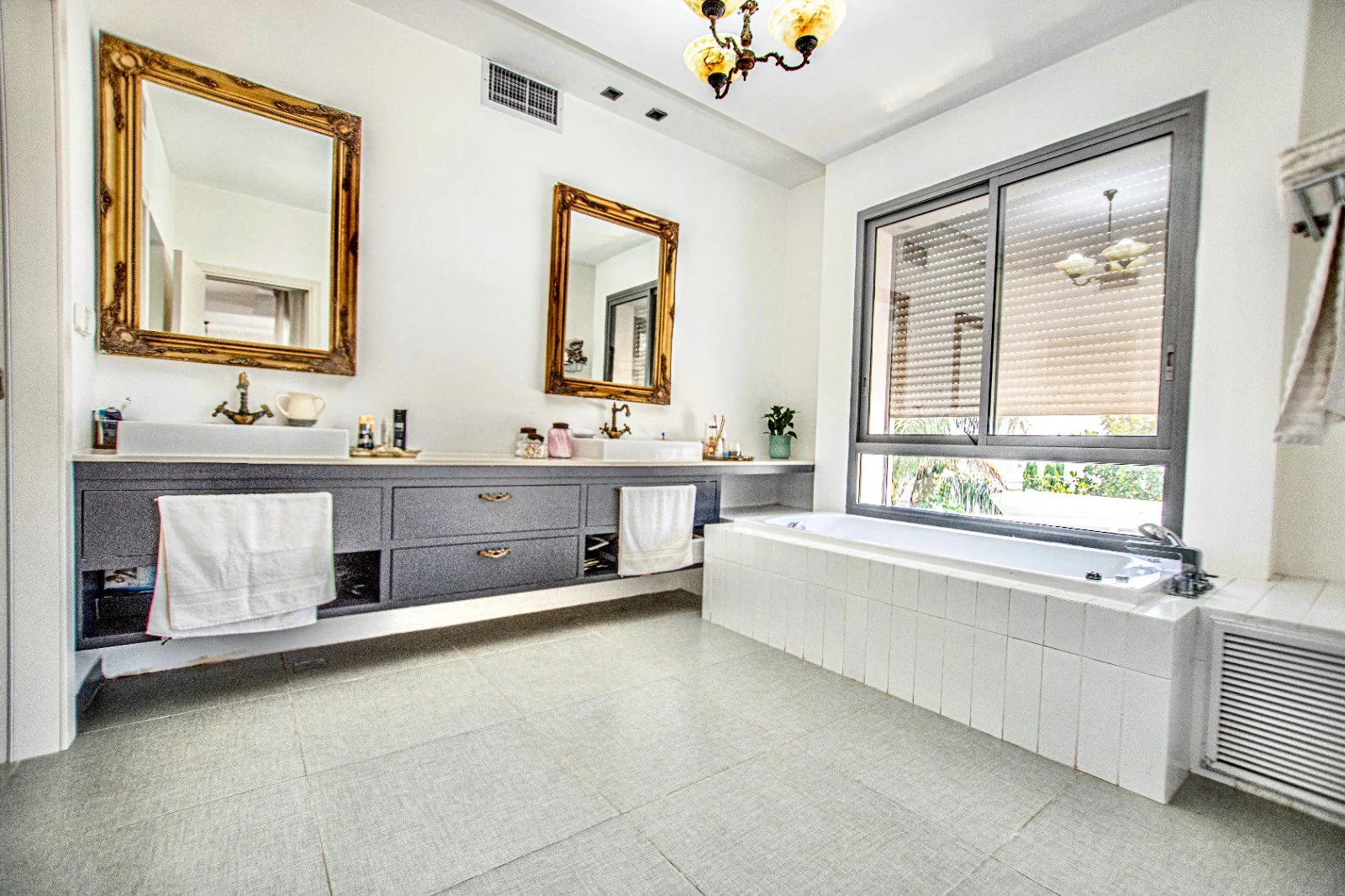Bathroom with two sinks and mirrors, gray cabinet, large window with blinds, bathtub, chandelier, and tiled floor.
