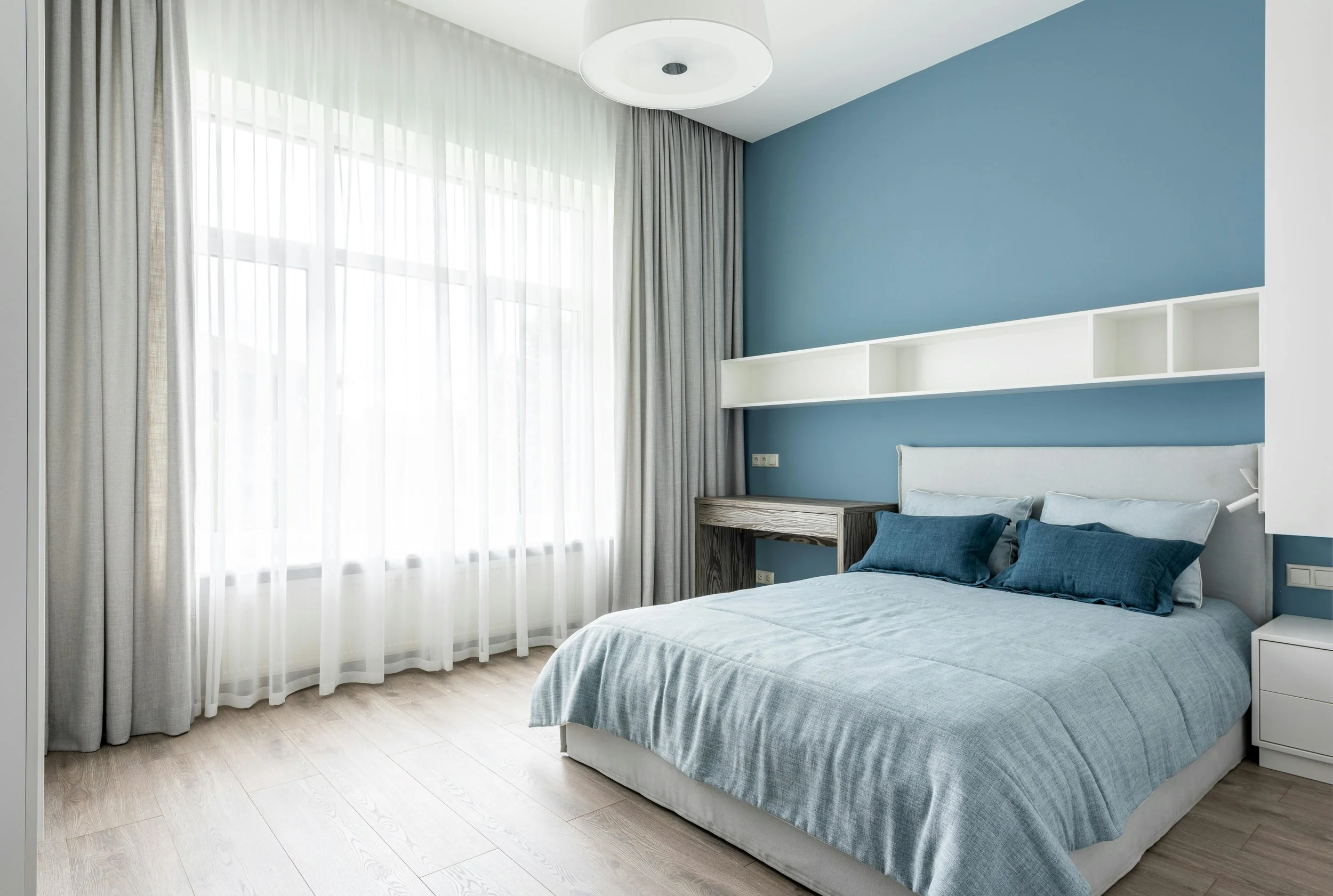 A bright bedroom with a large window with sheer white curtains and beige drapes. A bed with light blue bedding and two dark blue pillows is positioned against a blue accent wall. A white shelf is mounted above the bed, and a small wooden desk with a 