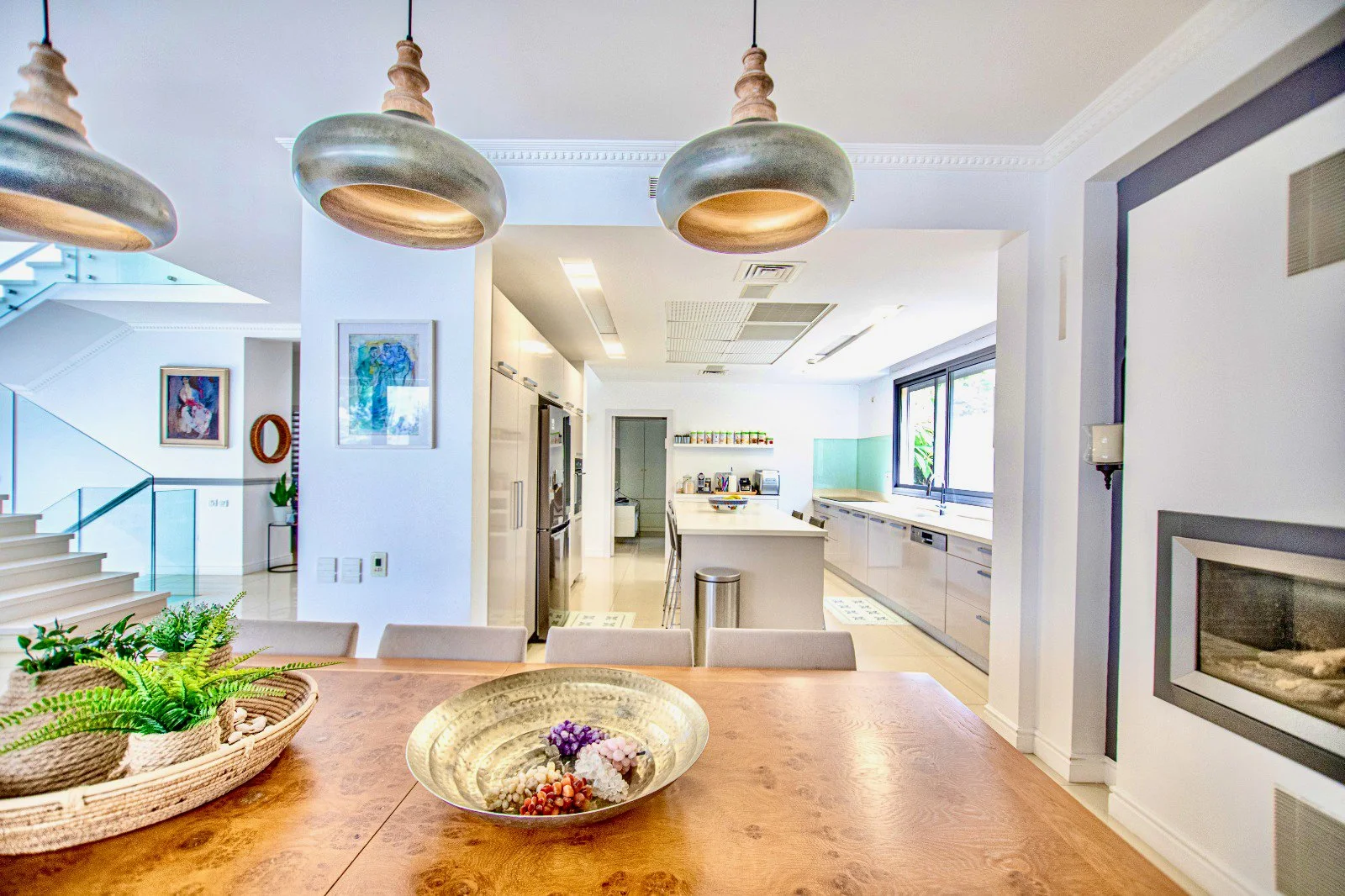 Spacious, modern kitchen with white cabinetry, large windows, stainless steel appliances, and a central island with bar stools. A wooden dining table with decorative items and greenery is in the foreground.
