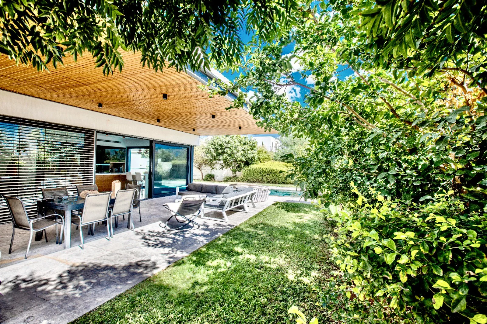 Modern house patio with outdoor dining table, chairs, lounge chairs, and a pool in the background, surrounded by greenery and trees under a partly cloudy sky.