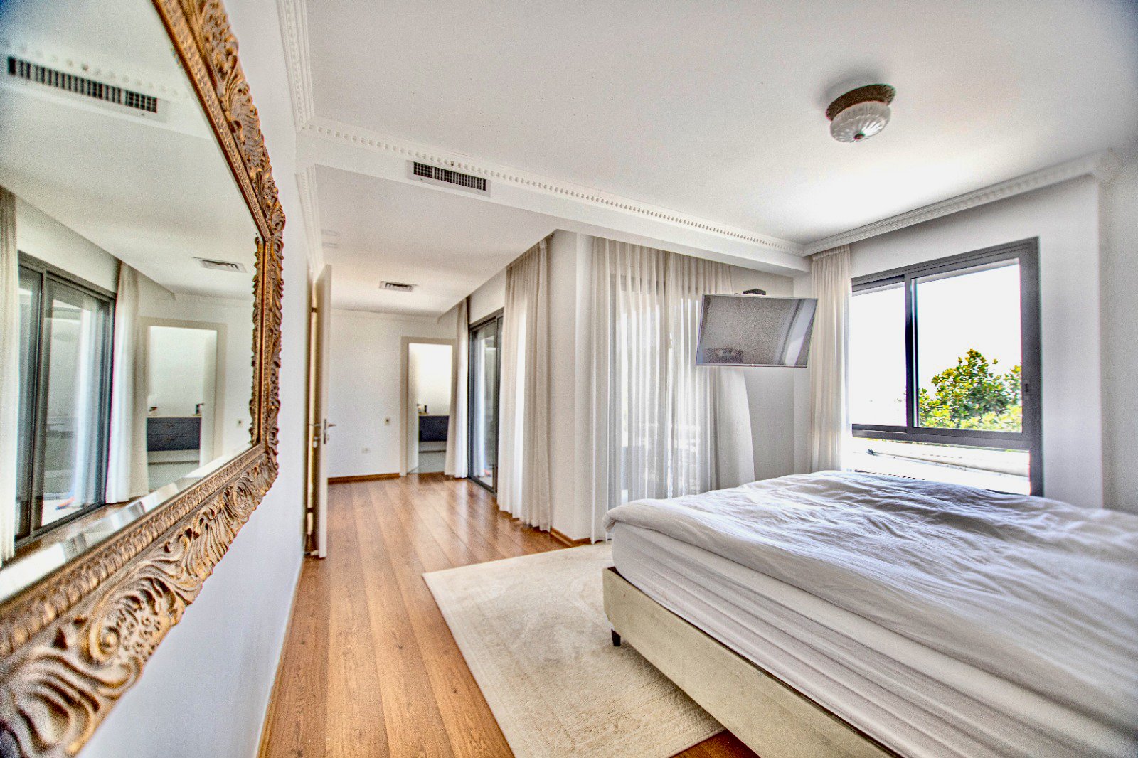 A bedroom with a bed, large window, wall-mounted TV, mirror with ornate frame, and wooden flooring.