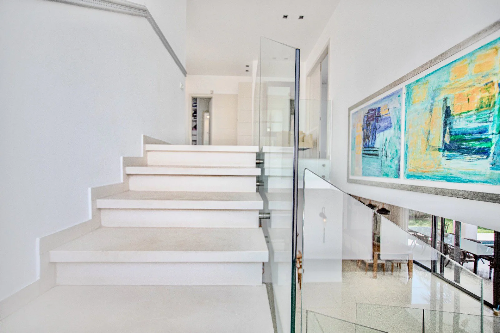 Interior view of a modern home staircase with white steps, glass railings, and contemporary wall art in the background.