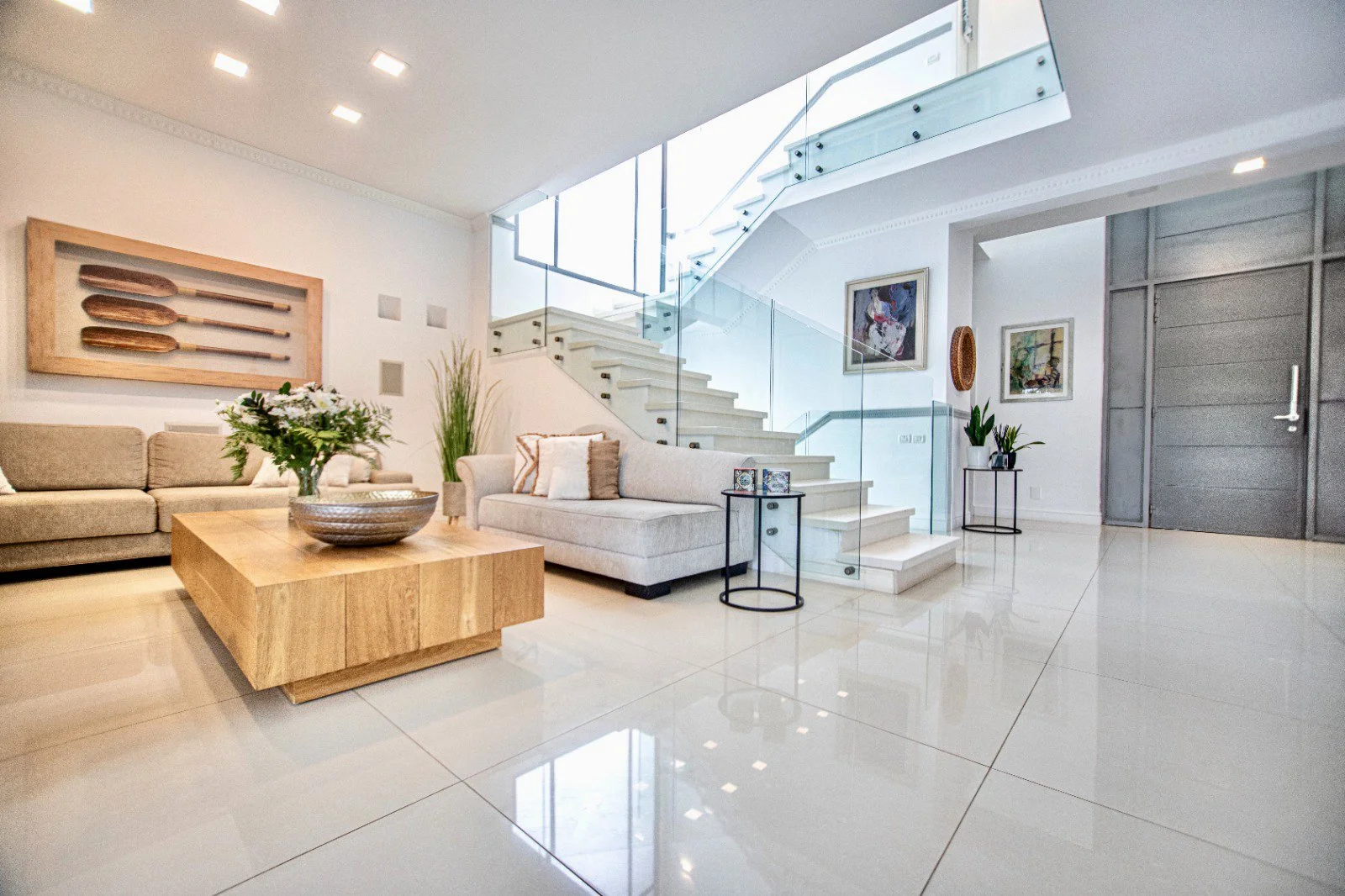 Modern living room with white and beige furniture, a large staircase with glass railing, and decorative elements including pictures on the wall and plants.