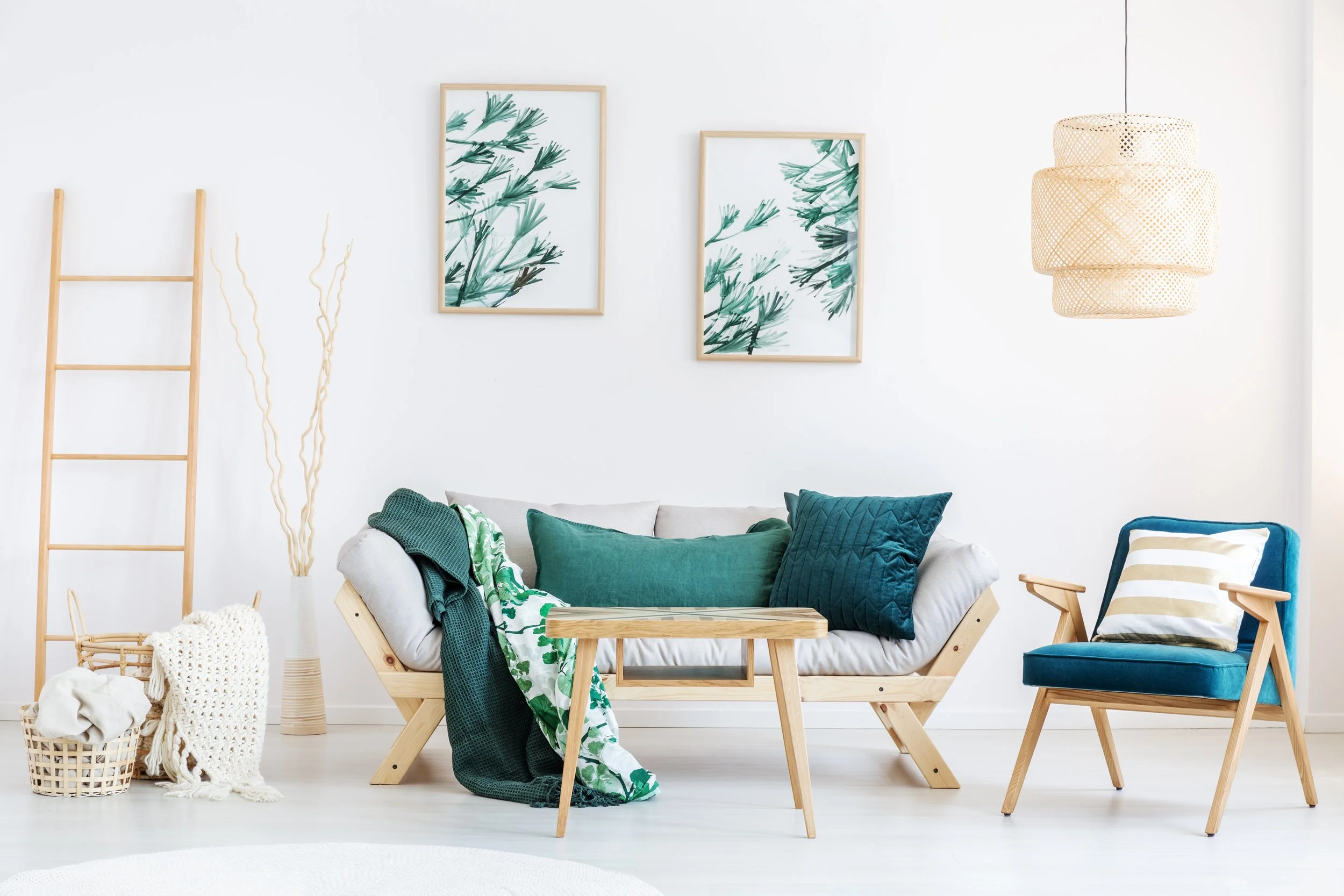 Bright living room with white walls, green framed botanical artwork, a light wood ladder, a white couch with green cushions and a dark green throw, a small light wood coffee table, a blue armchair with a white and beige striped pillow, a woven ceilin