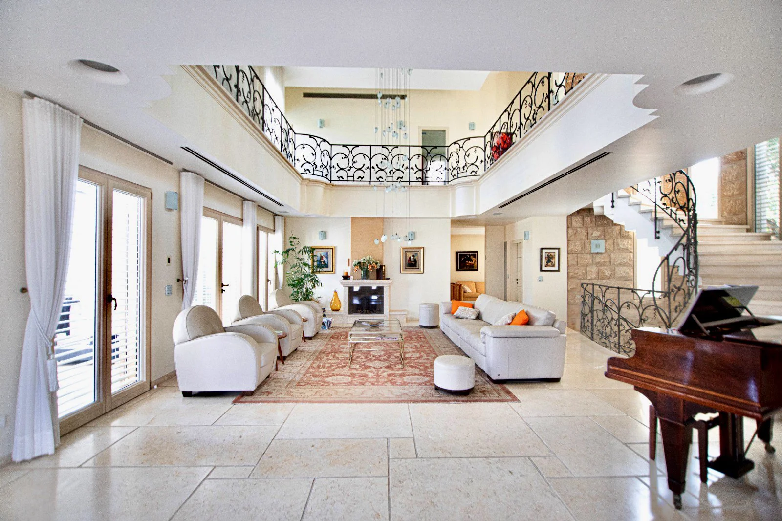 Spacious living room with white sofas, armchairs, and a grand staircase with ornate black iron railings, large windows with white curtains, and a fireplace with framed artwork above. There is a piano in the foreground and a patterned rug on the tile 