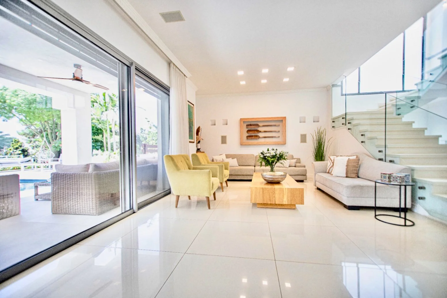 Modern living room with beige sofas, green armchairs, glass sliding door, white tiled floor, staircase with glass railings, and large windows letting in natural light.
