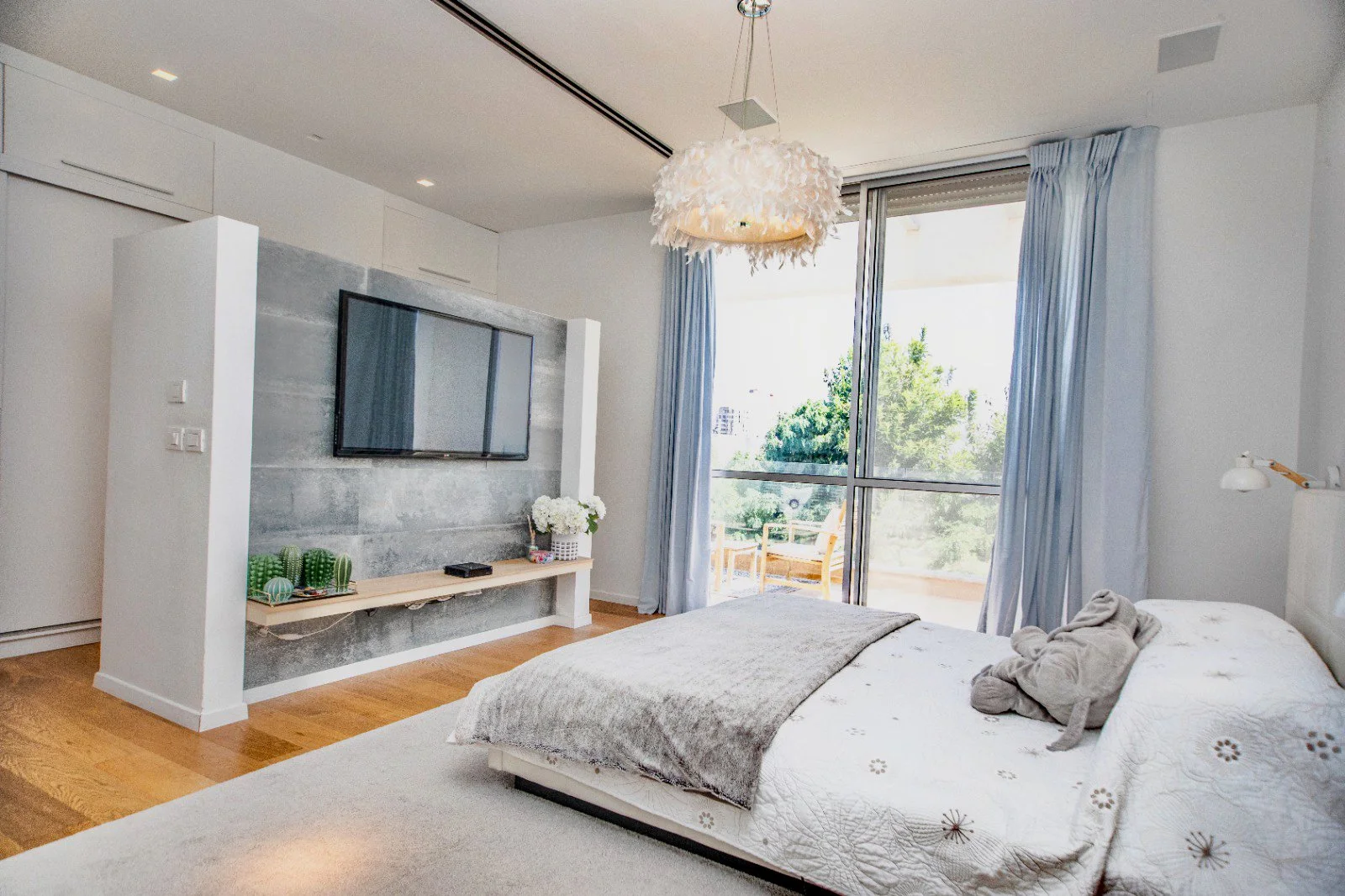 A bright bedroom with a large bed, light-colored bedding with a plush robe, a television mounted on the wall, a floating shelf with decorative cacti, flowers, a TV remote, a bubbly feathered ceiling light fixture, and a balcony with outdoor seating a