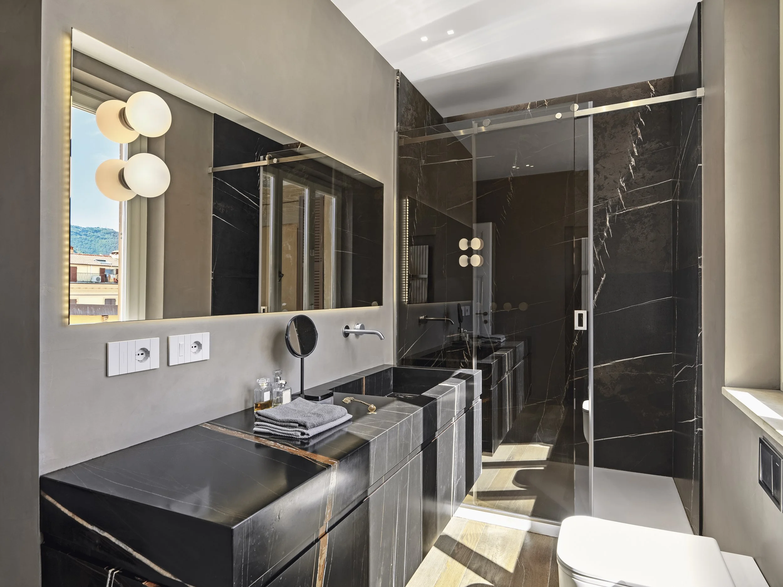 Modern bathroom with black marble countertops and walls, double sinks, large mirror, and walk-in shower with glass door, natural light from window, and wooden flooring.