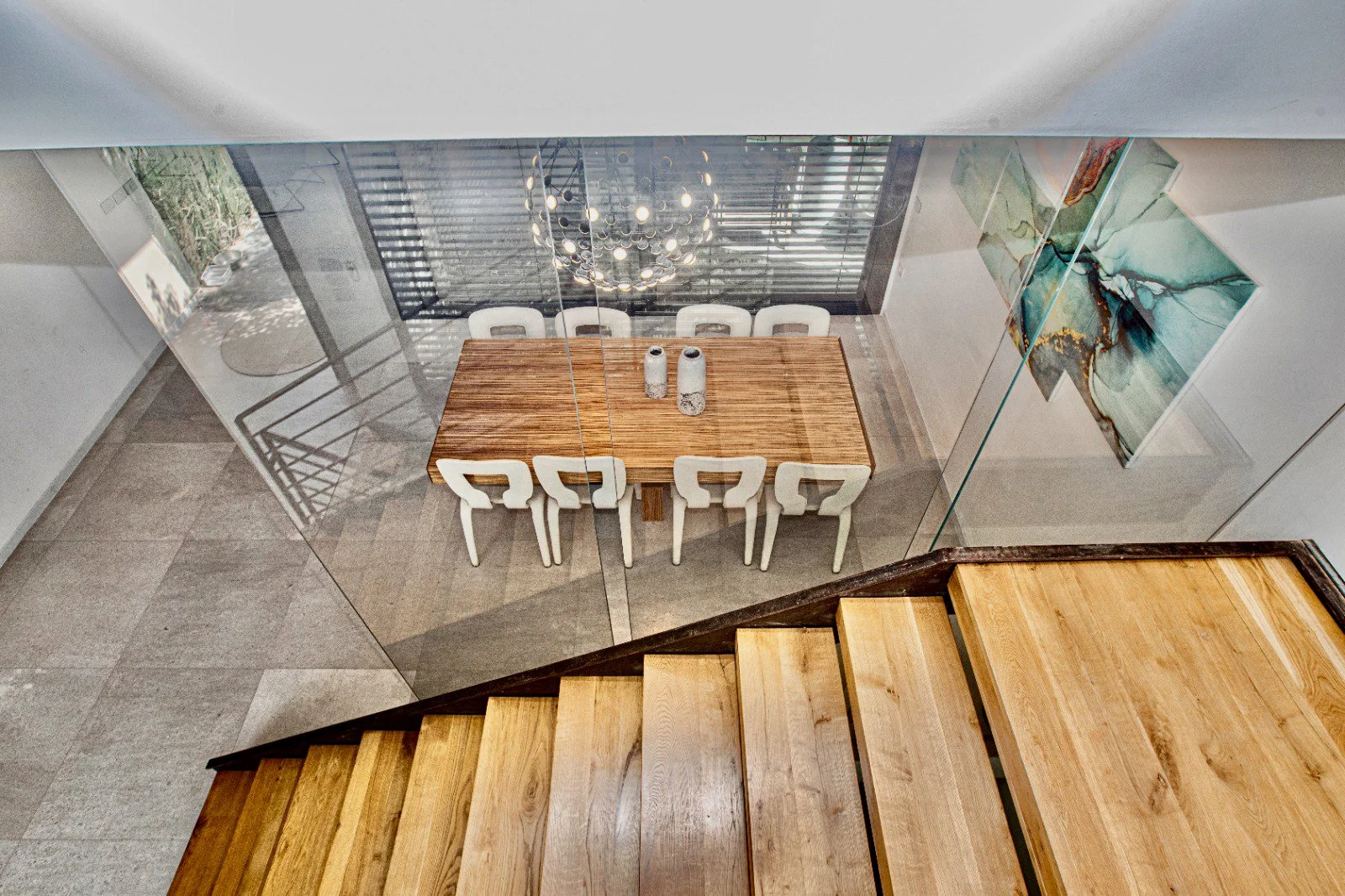 A dining room viewed from the staircase, featuring a wooden table with six white chairs, three decorative vases on the table, a modern chandelier hanging above, an abstract painting on the wall, and a large window with horizontal blinds and outdoor g