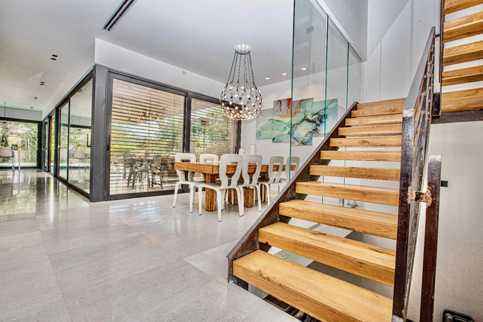 Interior of a modern house with a staircase, dining area, and glass walls with wooden blinds.