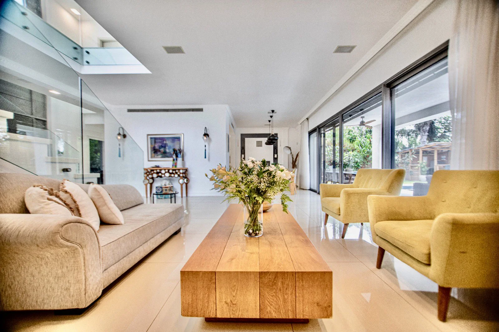 Bright living room with beige sofa, yellow armchairs, wood coffee table with floral arrangement, large sliding glass doors leading to patio.