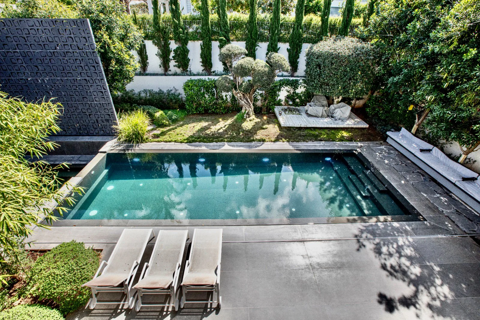 A backyard with a swimming pool, surrounded by lush green bushes and trees. There are four lounge chairs on the patio and a shaded seating area with chairs and a table in the background.