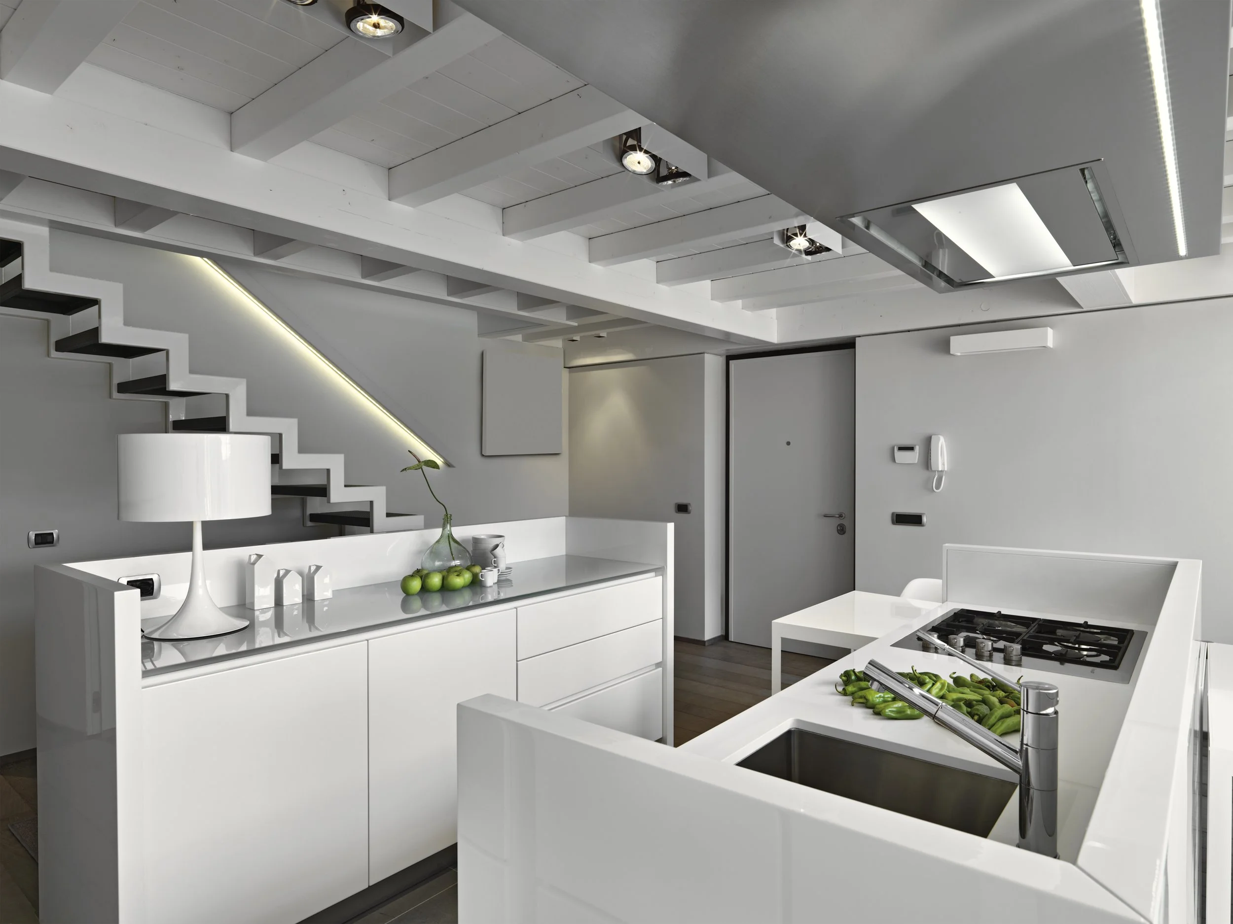 Modern kitchen with white cabinets, gray walls, and a kitchen island with a sink and stovetop. There are green bell peppers on the island, a white lamp, and a vase with green apples on the countertop. The ceiling has exposed beams and recessed lighti