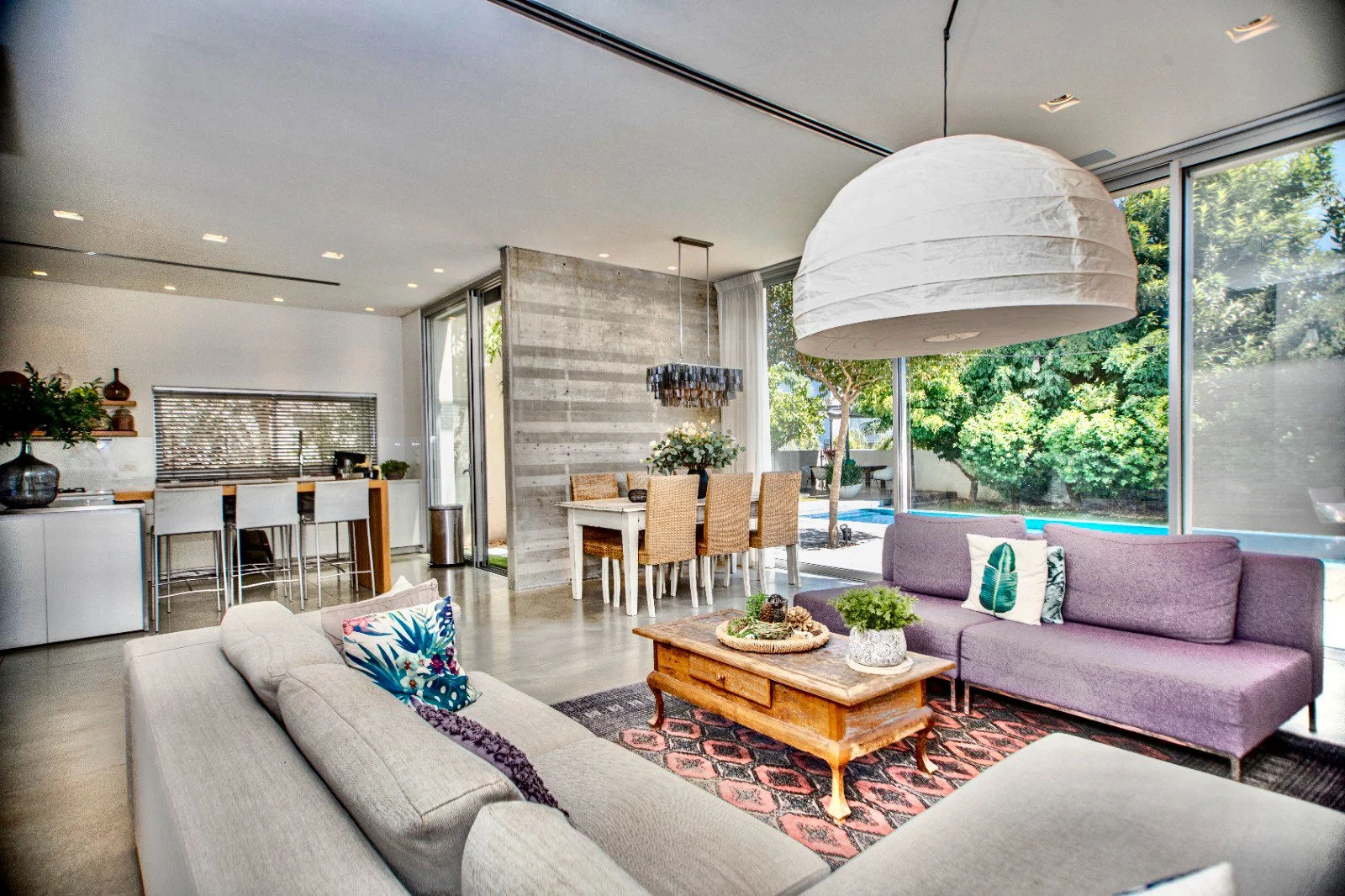 Modern living room with purple and beige sofas, a wooden coffee table with plants, a large white paper lantern pendant light, and large windows showing outdoor greenery.
